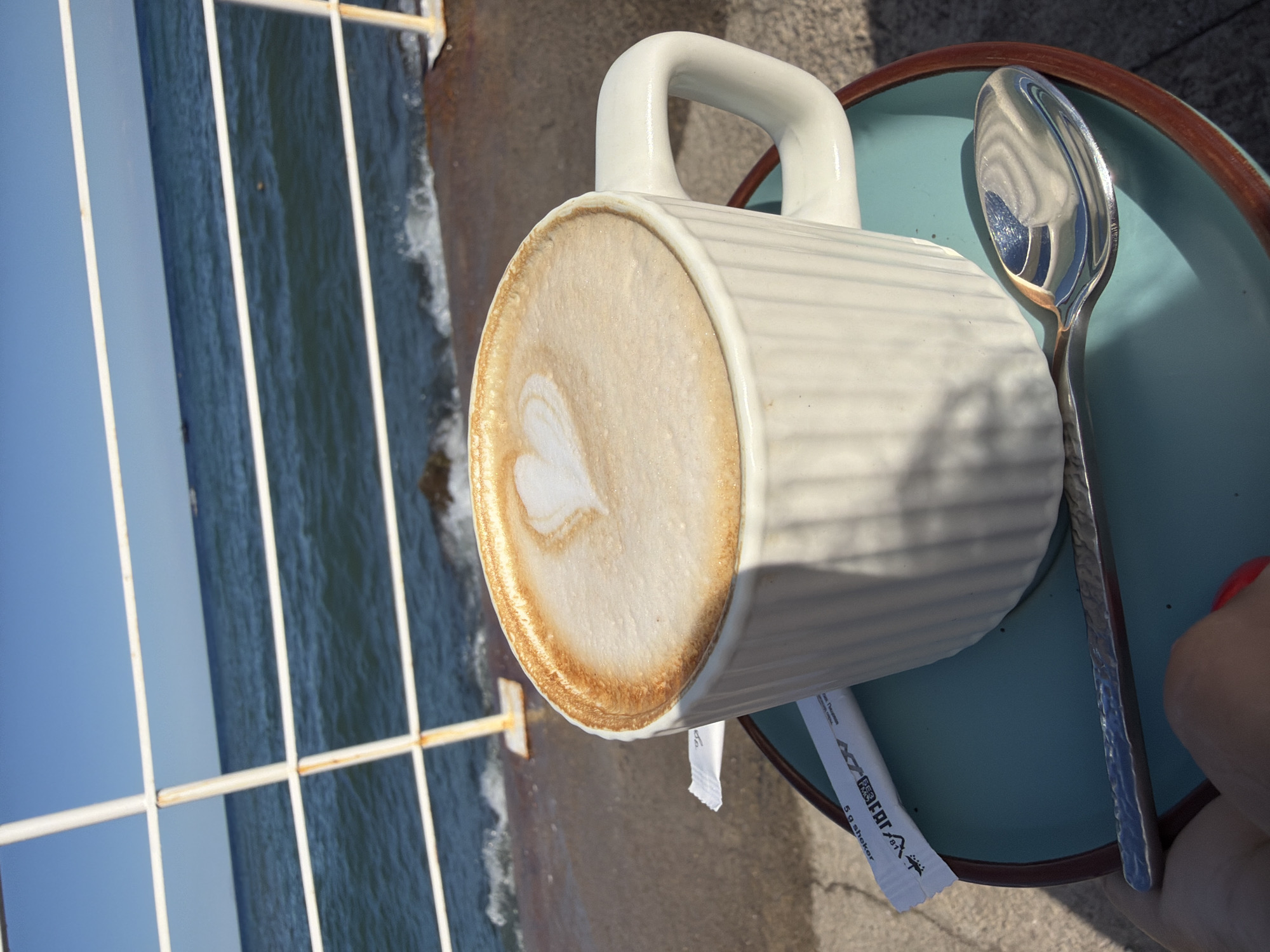 A white mug of coffee with latte art in the shape of a heart sits on a teal saucer. A silver spoon rests on the saucer, and a white packet of sweetener is next to the mug. The scene is outdoors, with a body of water, likely the Black Sea, in the background. A white metal railing separates the coffee from the water. The location is identified as Alushta, Ukraine. The sun is shining, casting shadows on the ground. A hand with a red fingernail is visible in the lower left corner, holding the saucer.