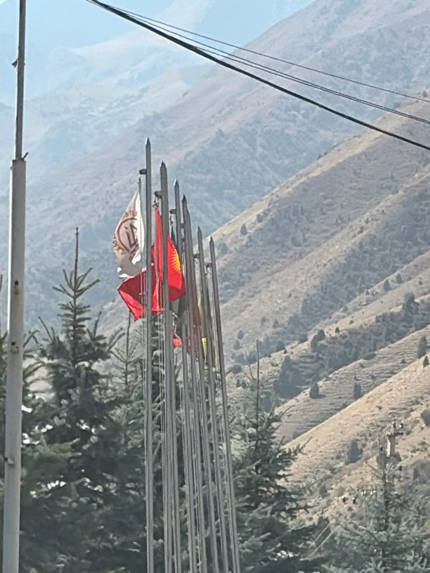 In an outdoor, daytime scene, a line of flagpoles stands tall in front of a backdrop of rugged, dry hills. The flags themselves are varied in design; one is white with brown lettering, another is red with a yellow sun emblem, and a third is partially visible and appears red. Evergreen trees are clustered in the foreground to the left, their branches dark against the lighter, arid landscape. Power lines stretch across the upper portion of the image, adding a modern element to the natural setting. The overall impression is one of a scenic, perhaps remote, location marked by both natural beauty and man-made structures.