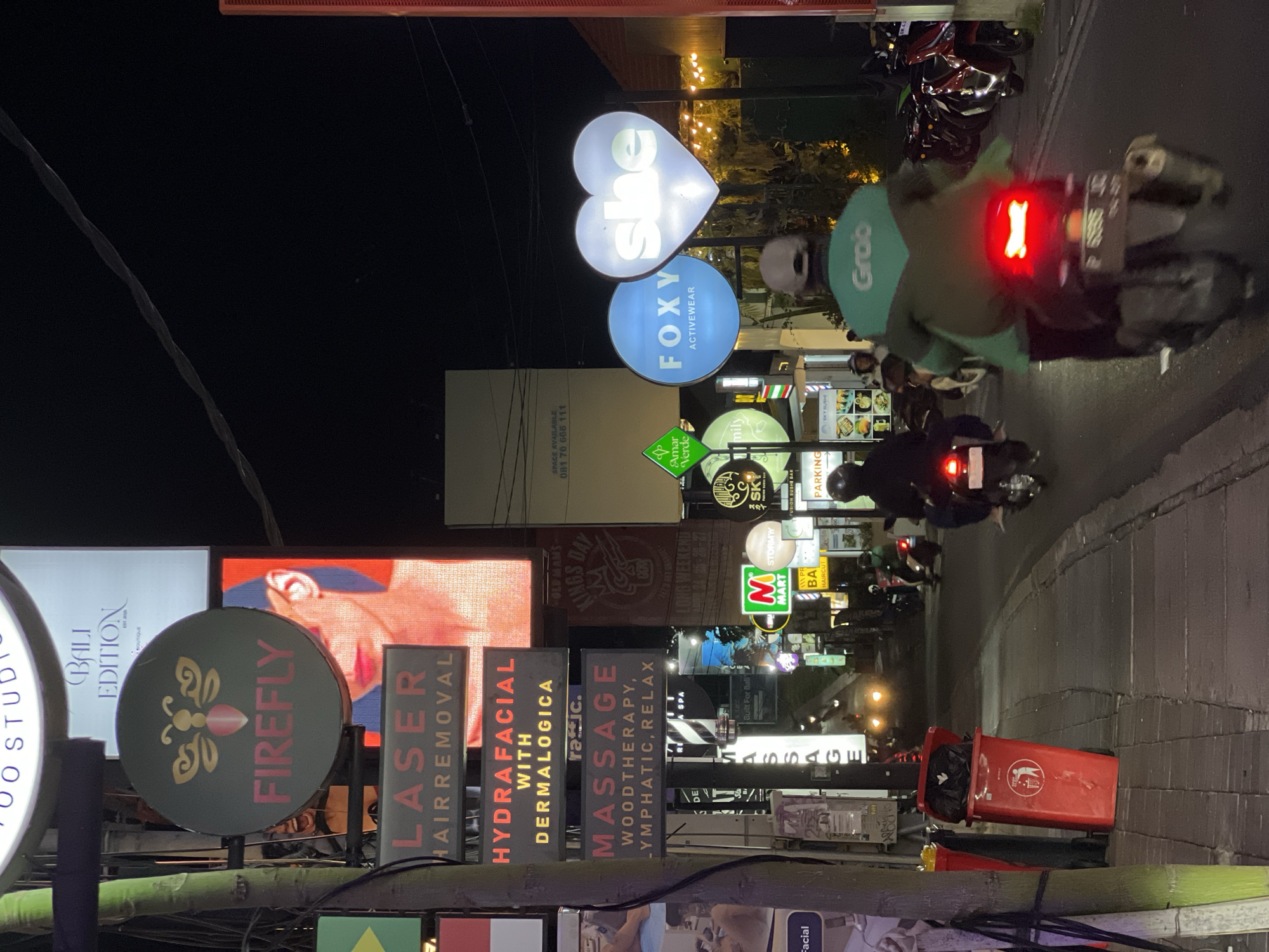 The image shows a street scene at night in North Kuta, Indonesia. The street is lined with illuminated signs for various businesses, including "Firefly," "Laser Hair Removal," "Hydrafacial," "Massage," "She," and "Foxy Activewear." There is also a "Grab" sign, indicating ride-sharing services. A red trash bin is visible in the foreground. Motorbikes are travelling down the street.