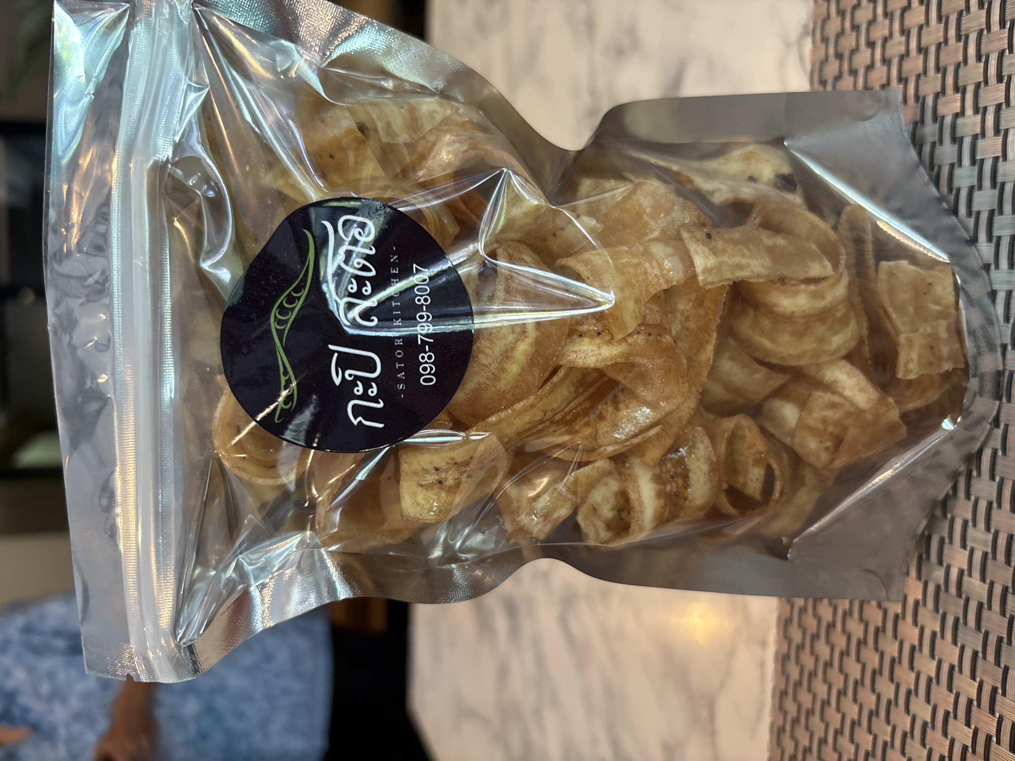 The image shows a sealed plastic bag of what appears to be banana chips, lying on a surface that resembles a table or countertop. The chips are golden brown and appear to be fried or baked. A black circular sticker is affixed to the front of the bag, containing text in Thai and English: "กะปิสะตอ - SATORI KITCHEN - 098-799-8007." The bag is reflective and filled with air, causing the chips to appear packed together. The background is slightly blurred, revealing a glimpse of another surface. The location is likely บ้านบางรักษ์, Thailand based on the Thai script on the sticker.