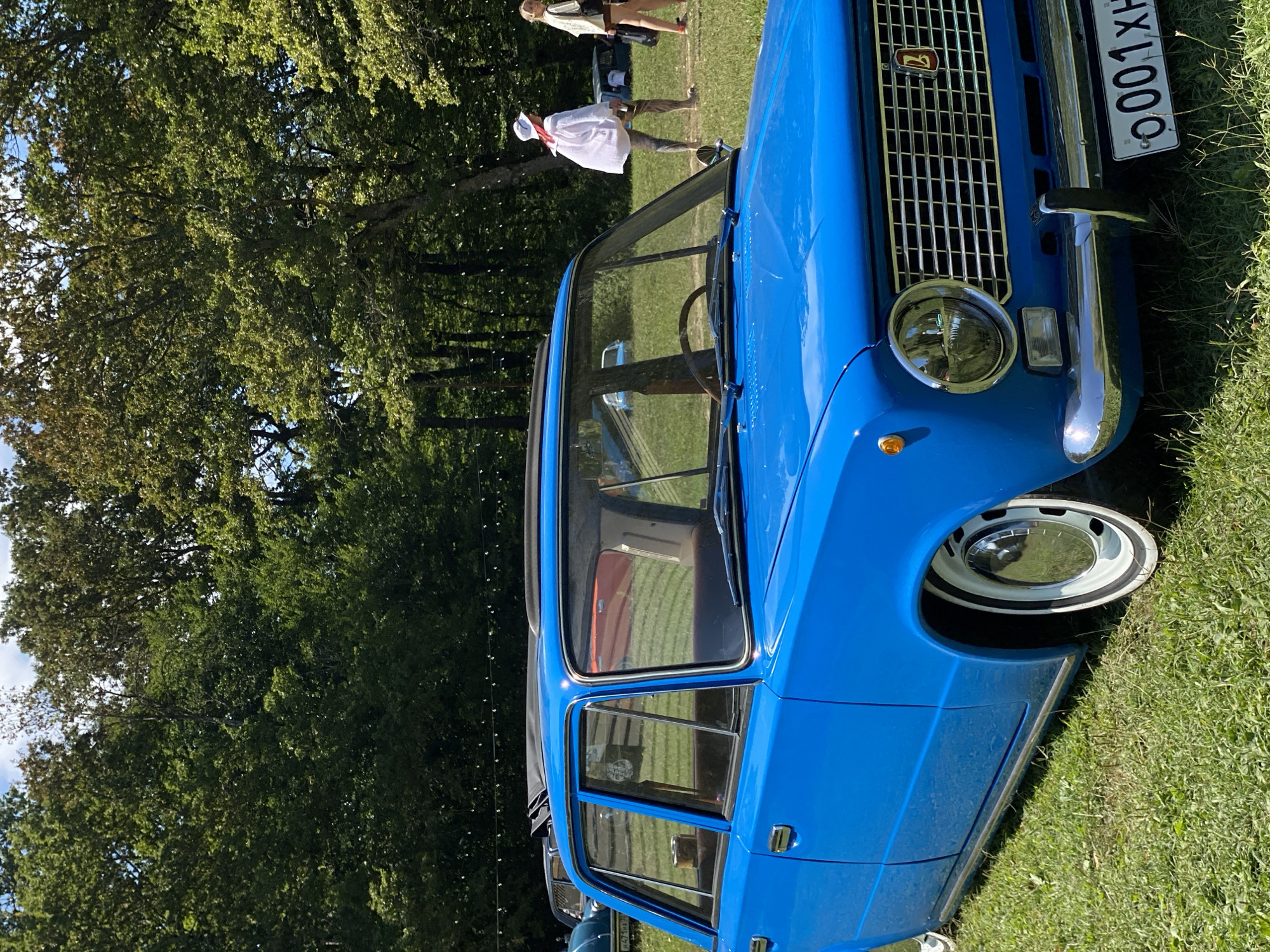 A bright blue, vintage sedan is parked on a grassy lawn in front of a dense line of trees. The car has a sun/moon roof and white-walled tires with chrome hubcaps. The front of the car features a chrome grille with a red logo and a license plate that reads "0001 XH". In the background, several people are walking across the grass, and other cars are visible parked amongst the trees. The overall scene suggests an outdoor event or gathering. The location is Bezymyannoe, Russia.
