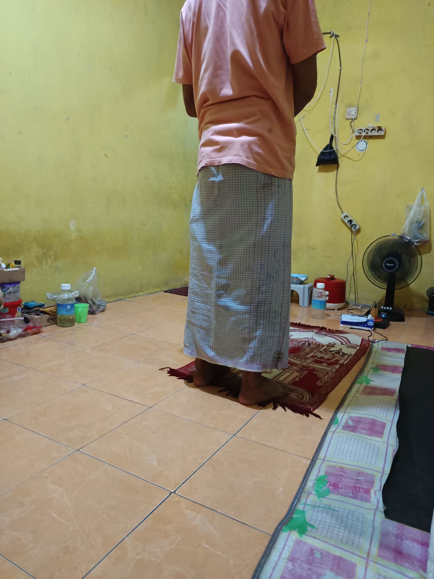The image shows a person standing on a prayer rug in what appears to be a room in Talaga Sari, Indonesia. The room has light yellow walls and a tiled floor. In the background, there are electrical outlets, wires, a fan, and various objects on shelves and the floor, including bottles and containers. The person is wearing a light-colored t-shirt and a patterned sarong. They are facing away from the camera, in a prayer posture.