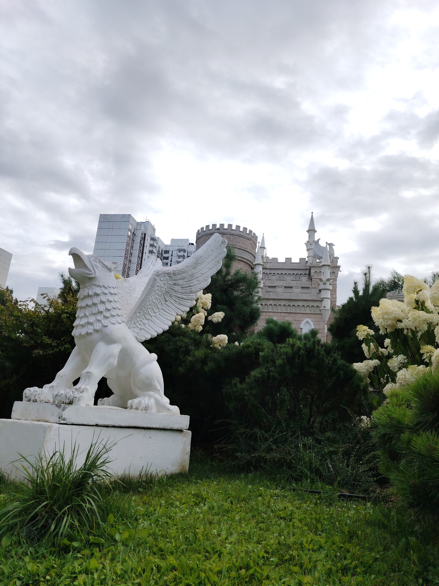The image depicts a white griffin statue in the foreground, positioned on a pedestal in a grassy area. The statue is detailed, with outstretched wings and an open beak. Behind the statue, a cluster of dark green trees and shrubs are visible, interspersed with white flowers. In the middle ground, a light-colored, fairy-tale-like castle with turrets and battlements stands amidst the greenery. To the left of the castle and behind the trees, several tall, modern apartment buildings are visible. The sky is overcast with gray clouds. The scene is located in the city of Zelenograd, Russia, specifically in the vicinity of 13 Serebryanaya Ulitsa, a residential area known for its unique architectural elements.