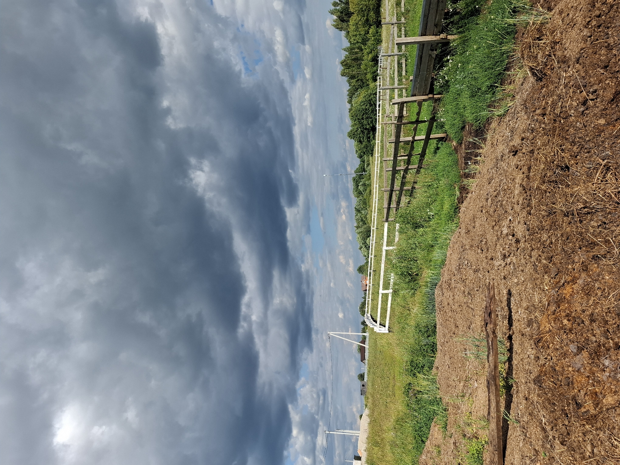 The image depicts a rural landscape under a heavily clouded sky, likely on the outskirts of Косяково, Russia. In the foreground, a large pile of dark brown organic material, possibly manure or compost, lies on the ground. A weathered wooden beam rests horizontally across this pile. Beyond the compost, a green field stretches out, interspersed with patches of tall grass and wildflowers. A wooden fence with vertical posts and horizontal rails runs through the middle ground, separating the field from a further area. To the left of the wooden fence, a white railing fence is visible. In the background, a dense line of green trees forms a treeline. Several power poles with wires are scattered across the horizon, and various buildings, including what appear to be houses with red roofs, are visible among the trees. The sky is dominated by dark, heavy clouds, with some lighter patches of blue sky and white clouds visible towards the horizon. The overall impression is one of a somewhat overcast day in a rural setting.