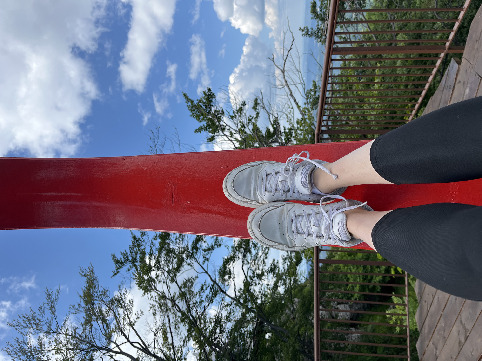 The media file shows a close-up, low-angle view of a person's legs and feet, wearing white athletic shoes and black leggings. The person is sitting on a bright red, flat surface, which appears to be part of a structure, possibly a bench or a platform. This red surface extends diagonally across the frame.
Beyond the red surface, the scene transitions to a view of trees, foliage, and a wooden railing on the right side, suggesting an outdoor setting like a park, a viewpoint, or a balcony. The background reveals a bright blue sky with scattered white clouds, indicating it is daytime. The lighting suggests it is a sunny day, possibly late morning or afternoon. The trees are green and leafy, with some bare branches visible, hinting at a temperate climate. The wooden railing has a rustic appearance.
The image composition focuses on the person's attire and the immediate surroundings, creating a sense of casualness and relaxation. There are no discernible emotions conveyed, but the context suggests a moment of rest or observation in an outdoor scenic location. No text is visible in the media file.