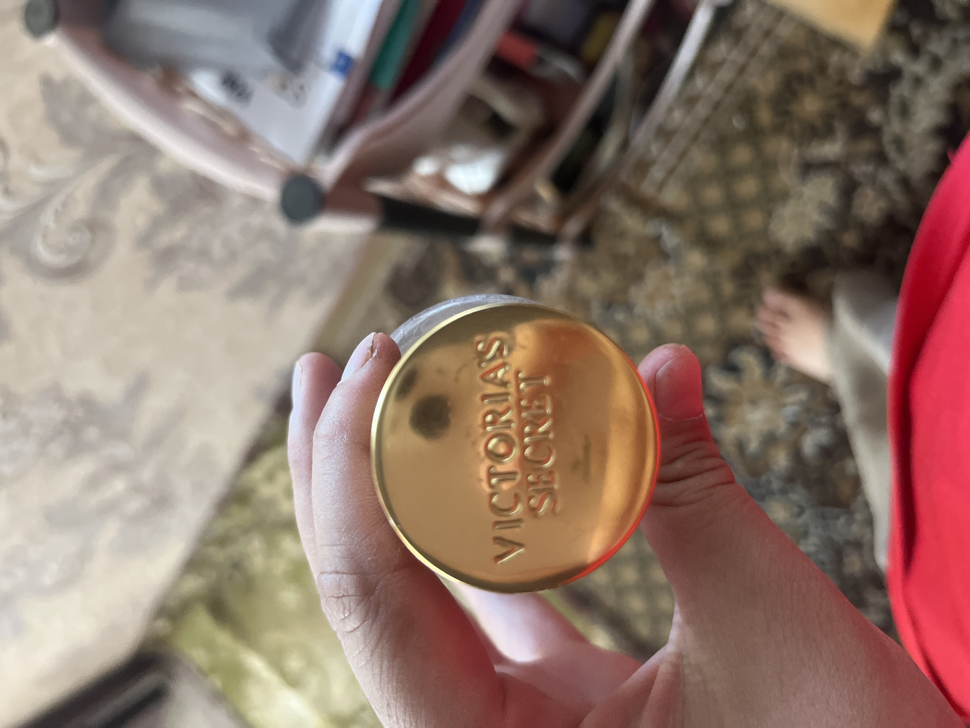 A hand holds a gold-colored lid with the embossed text "VICTORIA'S SECRET". The lid is circular and appears to be part of a cosmetic product. In the background, a blurred image shows a patterned carpet, a foot, and a bag containing various items like pens and papers. The overall scene is indoors.