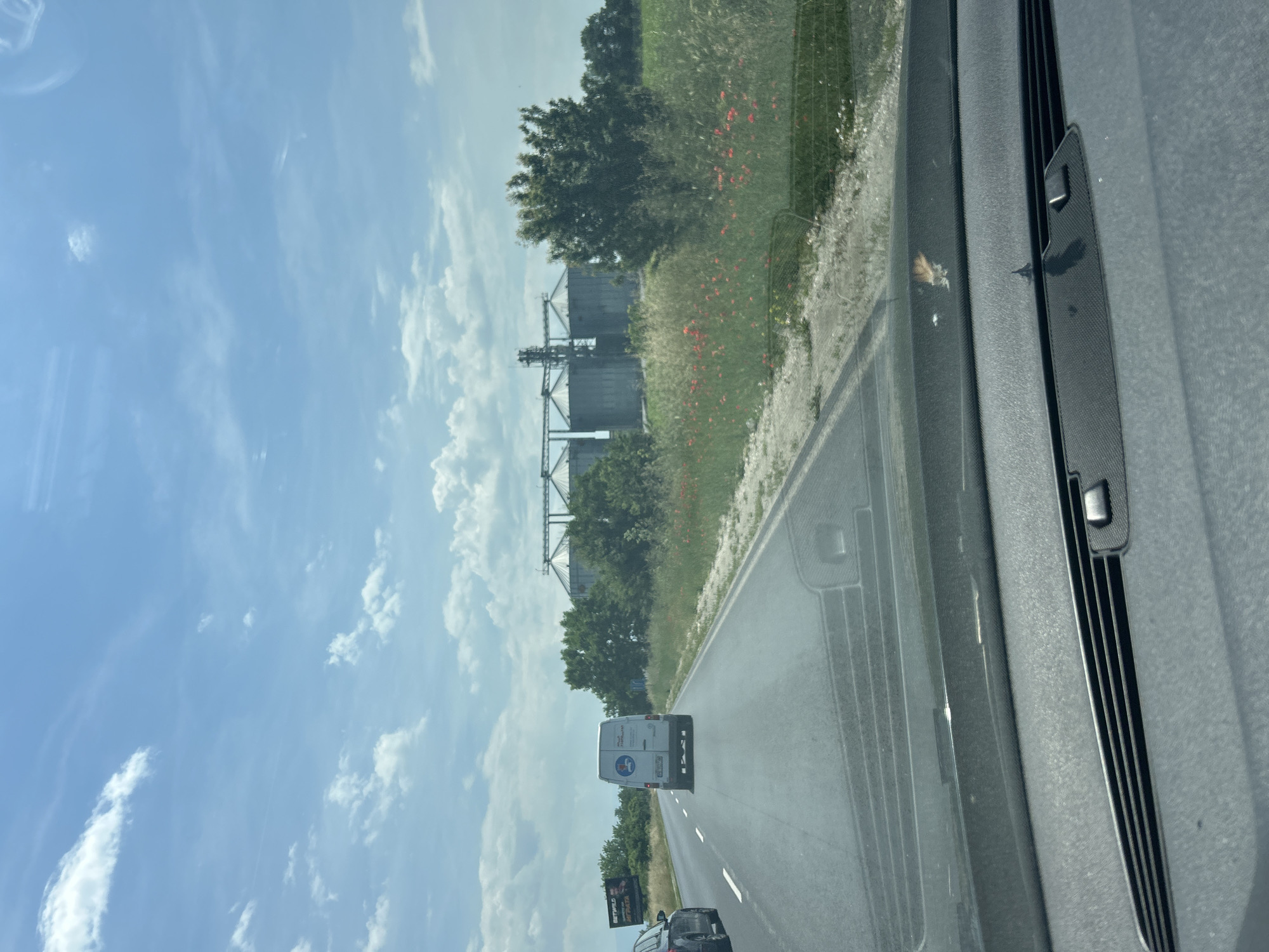 The image, taken from inside a moving vehicle, captures a daytime scene along a two-lane asphalt road, likely in Graf Ignatievo, Bulgaria. The sky is bright blue with scattered white cumulus clouds.
In the foreground, part of the car's interior, specifically the passenger-side dashboard and windshield frame with a wiper assembly, is visible. A small insect or piece of debris is present on the windshield.
On the road ahead, a white delivery van with a logo on its rear is visible. The logo depicts a stylized figure holding a package, with the word "RAPIDO" potentially visible below it, followed by smaller text. Behind the van, a dark-colored car is also traveling.
To the left of the road, a vibrant green landscape unfolds, featuring numerous trees and patches of bright red flowers, possibly poppies. Further in the distance, large, dark industrial silos, likely for grain storage, stand prominently against the sky, supported by intricate metal structures.
To the right of the road, a billboard is partially visible, clearly displaying the text "METRO" and "HIPERMARKET", indicating the presence of a supermarket nearby. The overall scene suggests a journey through a mixed agricultural and industrial area.