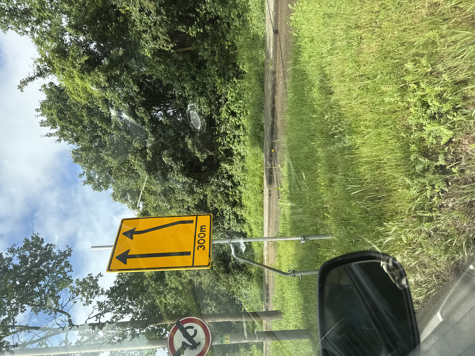 A bright daytime scene is captured from inside a vehicle, looking towards the right side of the road. The image is oriented vertically, but if rotated to landscape, it shows a road setting with lush green trees lining the left and a strip of green grass bordering a concrete edge on the right. The sky, with patches of blue and white clouds, is visible through the tree canopy.
In the mid-ground, a prominent yellow rectangular road sign is displayed. It features two black parallel arrows pointing forward and slightly upwards, suggesting a lane merge or traffic direction. The text "300m" is clearly visible on the right side of the sign, indicating a distance. A small "BUKO" label is also present on its lower edge. The sign is mounted on metal poles.
Below and to the right of the yellow sign, a circular road sign with a red border and white background is partially visible, though its central symbol is obscured. Faint text, "CEI R" and "177", can be seen on its support structure. Part of the vehicle's side mirror is visible in the bottom right corner of the frame. The overall impression is a sunny day on a road in a verdant, possibly rural, area.