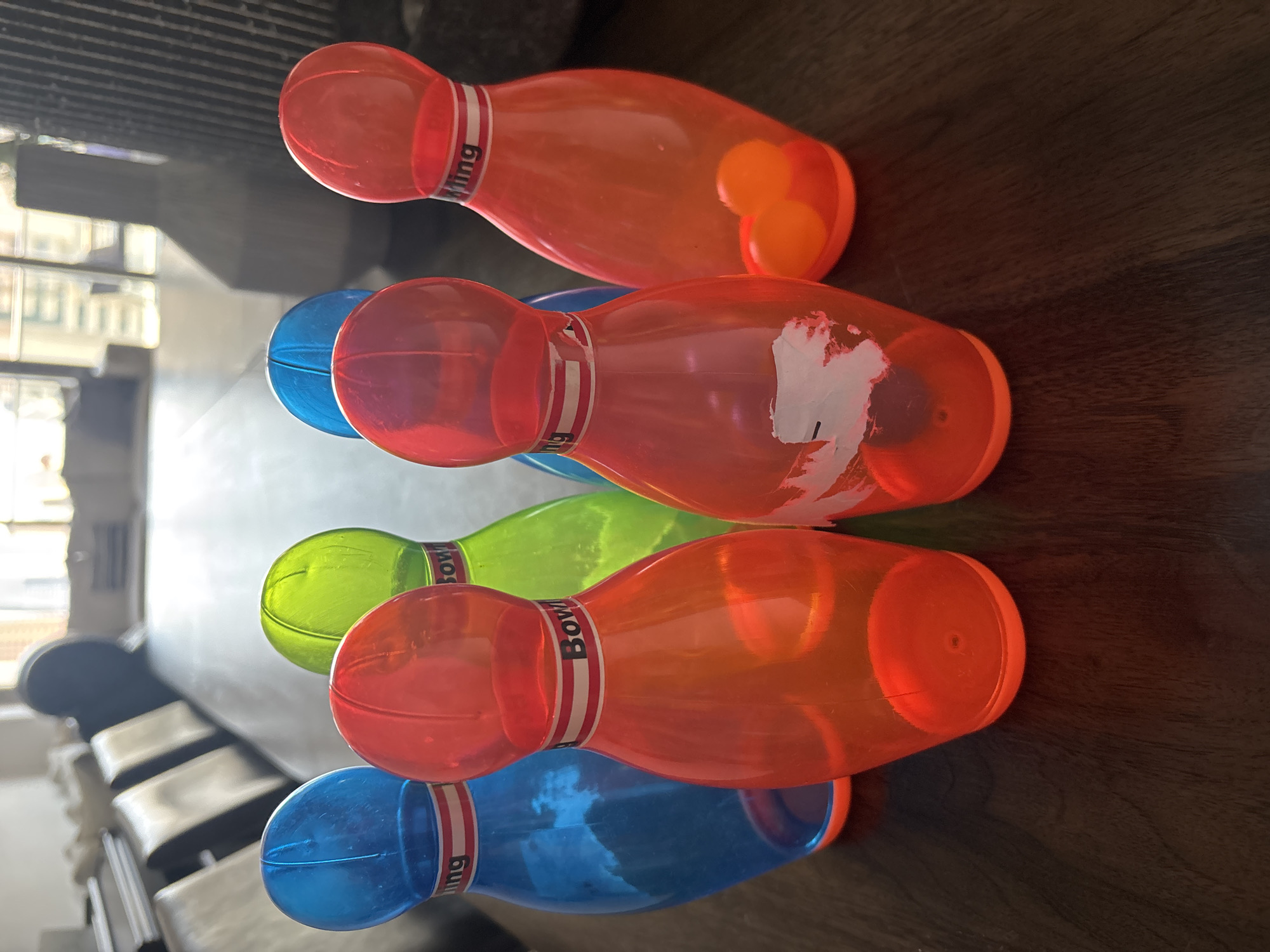 The image displays a set of six plastic bowling pins, three red, two blue, and one green, arranged on a dark wooden surface. Two small orange balls are visible inside one of the red pins. The pins are translucent and appear to be toys, as suggested by their bright colors and size. The background is blurred, showing what appears to be a dining room with chairs and windows, suggesting an indoor setting. There is no discernible information in the image to place it in Dubai, United Arab Emirates, or any other specific city.