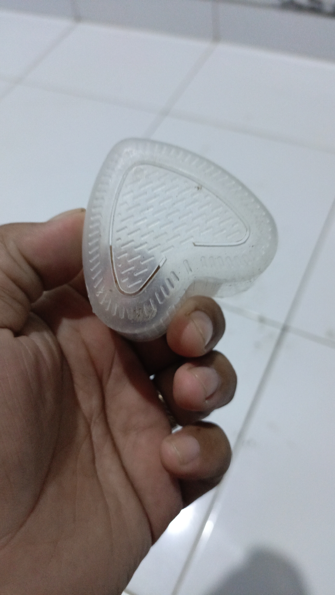 A person's hand is holding a translucent, triangular-shaped plastic mold with a textured interior, against a tiled floor background. The mold is positioned at an angle, with the textured side facing upwards. The hand is brown-skinned and appears to be that of an adult. The floor is made of white square tiles. The scene is indoors, likely a residential or commercial space. The location is identified as Trihanggo, Indonesia.