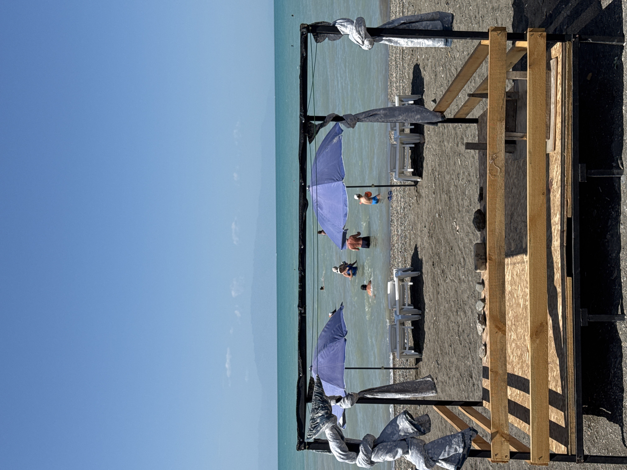 The image depicts a beachfront scene under a clear blue sky with a distant mountainous horizon. In the foreground, a rustic, unfinished wooden structure with a metal frame occupies the lower portion of the frame. Two large, light purple beach umbrellas are set up on the dark sand beach, each accompanied by a white lounge chair. Several people are visible in the shallow, turquoise water, some swimming and one person holding an orange object. Two pieces of patterned fabric are draped over the vertical supports of the foreground structure. The environment suggests a sunny day at a beach resort. The image does not contain enough visual information to confirm its location in Zharbulak, Kazakhstan.