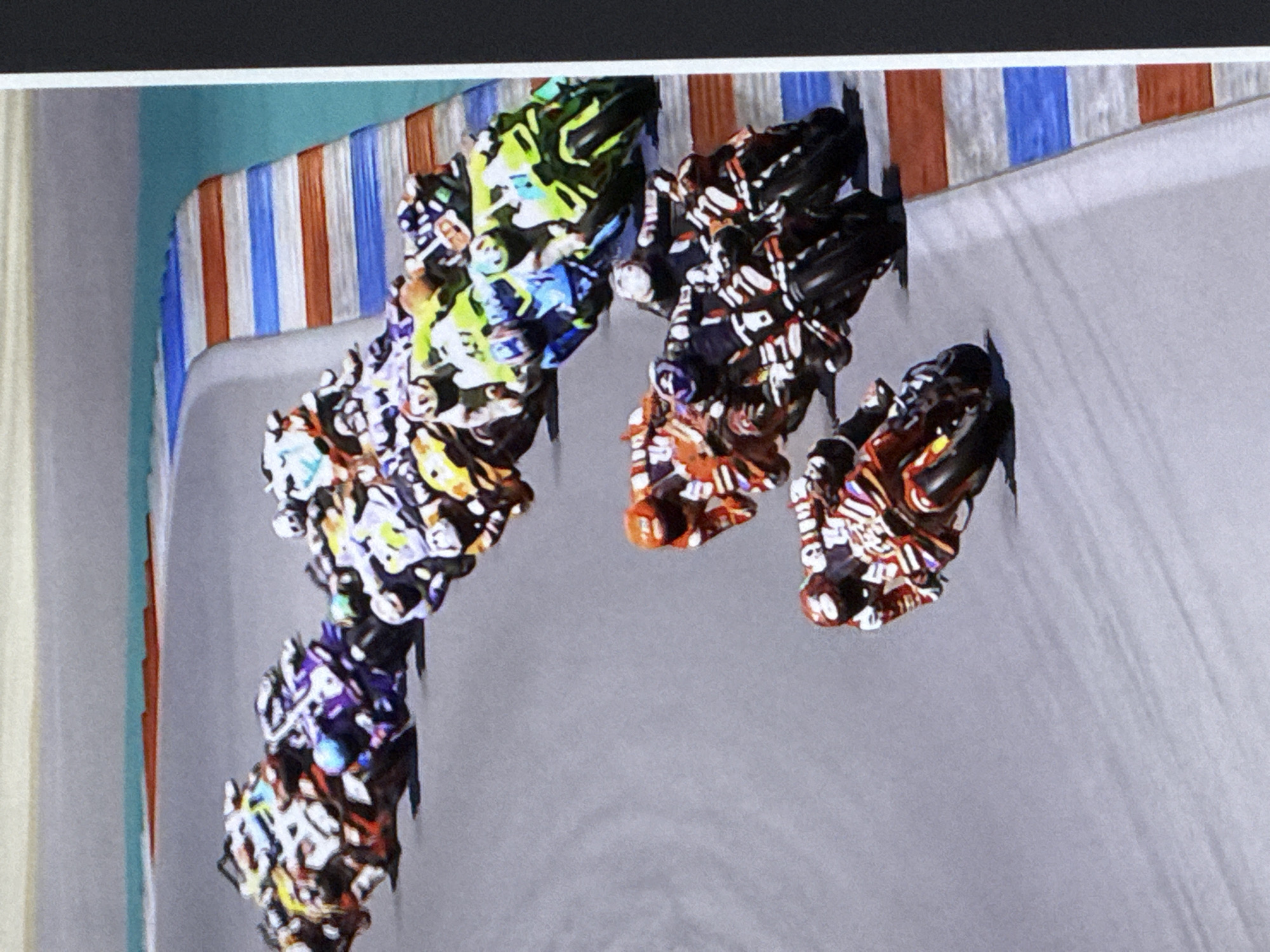 The image depicts a high-angle view of a motorcycle race, likely a video game simulation or a photograph of one. Multiple motorcycles, ridden by people in colorful racing suits, are tightly bunched together on a grey asphalt track with a blue and orange striped curb on the right side, bordering a teal body of water. The motorcycles are at various angles, suggesting they are navigating a turn. The focus is on the race action, with no discernible urban landscape or specific location in Cahul, Moldova visible. The environment appears to be a closed race track.