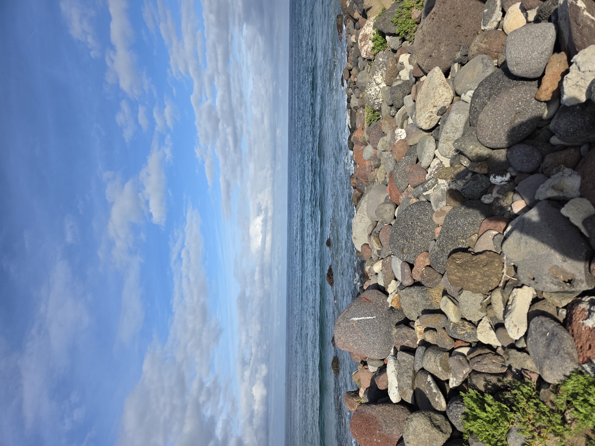 The image depicts a rocky shoreline meeting the ocean under a cloudy sky. The rocks are predominantly gray and reddish-brown, with some vegetation interspersed. The ocean stretches to the horizon, where it meets the sky. The location is likely near Ensenada, Mexico, given the coastal environment and rock formations. No people are visible in the image.