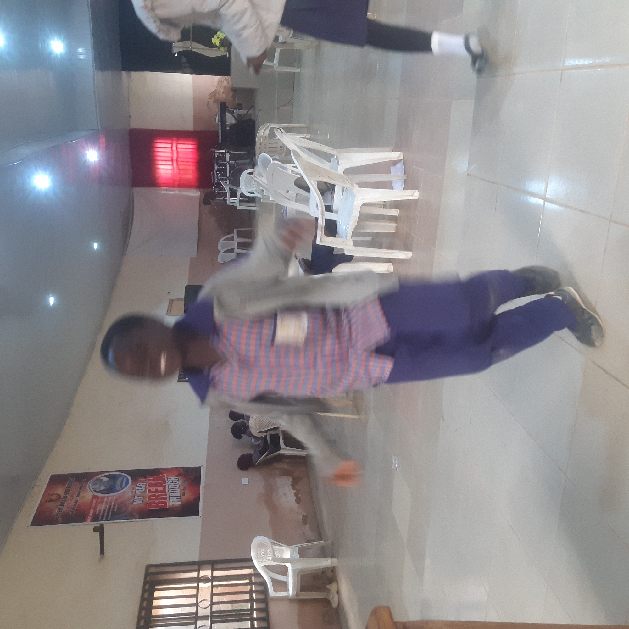 The image depicts an indoor scene, possibly a community hall or church, with beige walls and a tiled floor. In the foreground, a young person wearing a plaid shirt and blue pants is blurred due to motion, suggesting they are actively moving or dancing. A banner with the text "MY YEAR OF BREAK THROUGH" is visible on the back wall. Several white plastic chairs are scattered throughout the room. In the background, other individuals are seated or standing, some of whom are also out of focus. Musical instruments, including drums and a keyboard, are set up on a raised platform. The lighting appears to be artificial, with overhead lights illuminating the space.