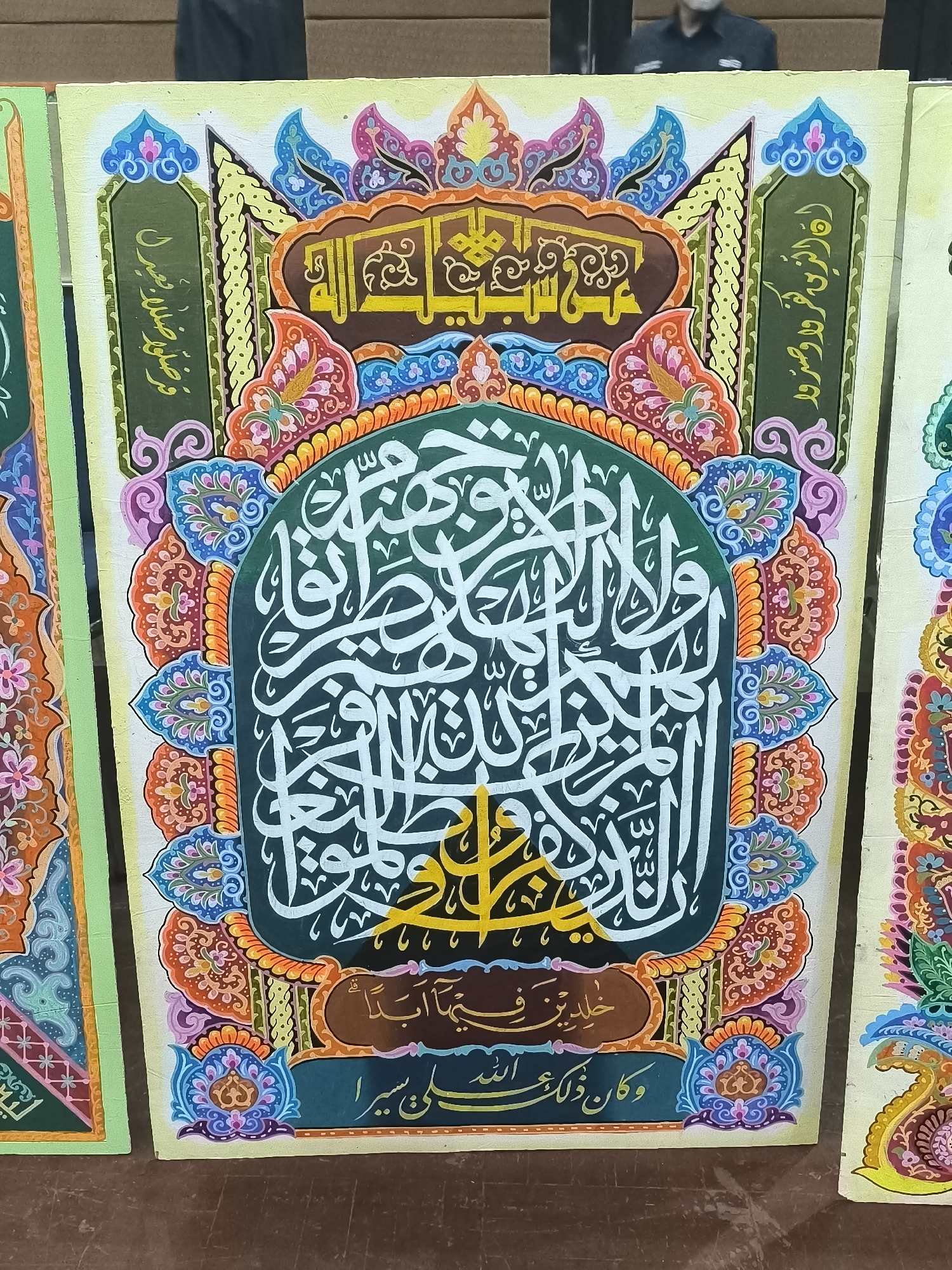 A close-up, head-on shot showcases a vibrant Islamic calligraphy artwork displayed on a wooden surface. The artwork is an intricate composition of Arabic script rendered in white against a dark green background, framed by a circular, elaborate border of colorful floral and geometric patterns in shades of orange, blue, pink, and yellow. The outermost layer features a yellow frame with darker yellow decorative elements. The script itself is densely packed, with some larger, stylized characters. Below the central calligraphy, there is a smaller brown panel with more Arabic text, followed by another line of white script on a dark green background. The overall aesthetic is rich and decorative. The context of Minasa Te'ne, Indonesia, is not discernible from the visual information provided in the image.