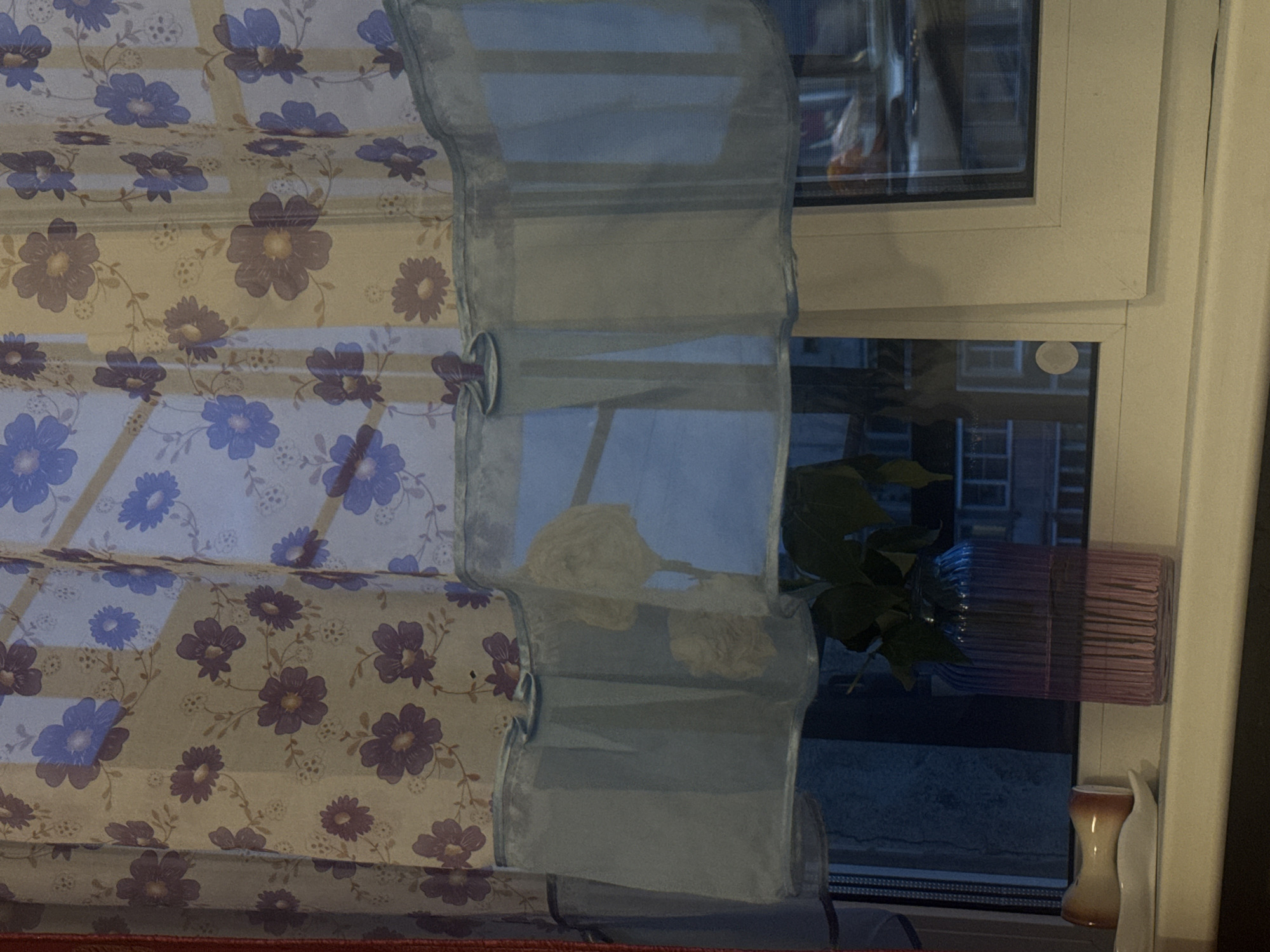 The image depicts a window in an apartment, likely in an urban setting in Russia, as suggested by the architectural style visible through the glass. The window is adorned with a sheer curtain featuring a floral pattern of purple and blue flowers. The lower portion of the curtain is a lighter blue, with a ruffled trim.
On the windowsill, there is a pink glass vase containing green leaves, suggesting a plant or cuttings. To the left of the vase, a small, beige and orange object, possibly a decorative piece or a spice grinder, rests on a white saucer.
The window offers a view of buildings outside, with visible windows and architectural details that are characteristic of residential buildings in Russia. The lighting suggests it is either dusk or evening, as the interior lights are not on, and the exterior light is dim. The overall scene conveys a sense of domestic interior, with a focus on the window and its immediate surroundings.