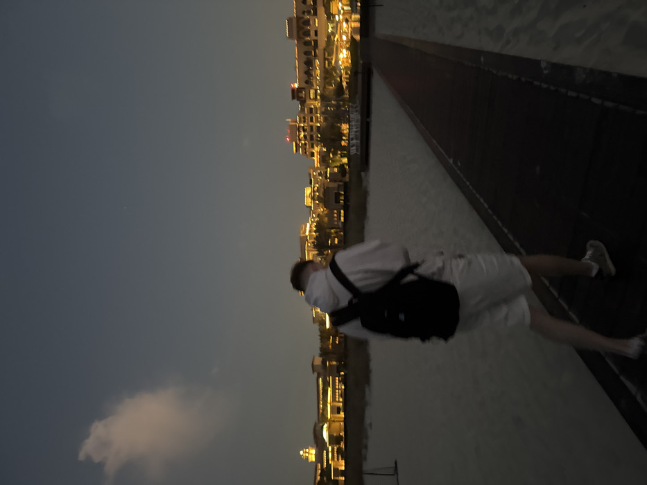 A male individual is depicted walking away from the camera along a dark wooden boardwalk on a sandy beach at dusk. He wears a white hooded sweatshirt, white shorts, and light-colored sneakers, and carries a black backpack. His hair is short and light-colored. The boardwalk extends into the middle distance, flanked by light-colored sand. In the background, a series of multi-story buildings, illuminated with warm yellow lights, line the horizon. These buildings feature distinctive architectural elements consistent with luxury resorts. The sky above is a dark blue-grey, with a single prominent white cloud in the upper left quadrant and a small, faint white point of light in the upper right. The scene is located on Saadiyat Island, Abu Dhabi, United Arab Emirates, consistent with the architecture of beachfront resort properties in that area.