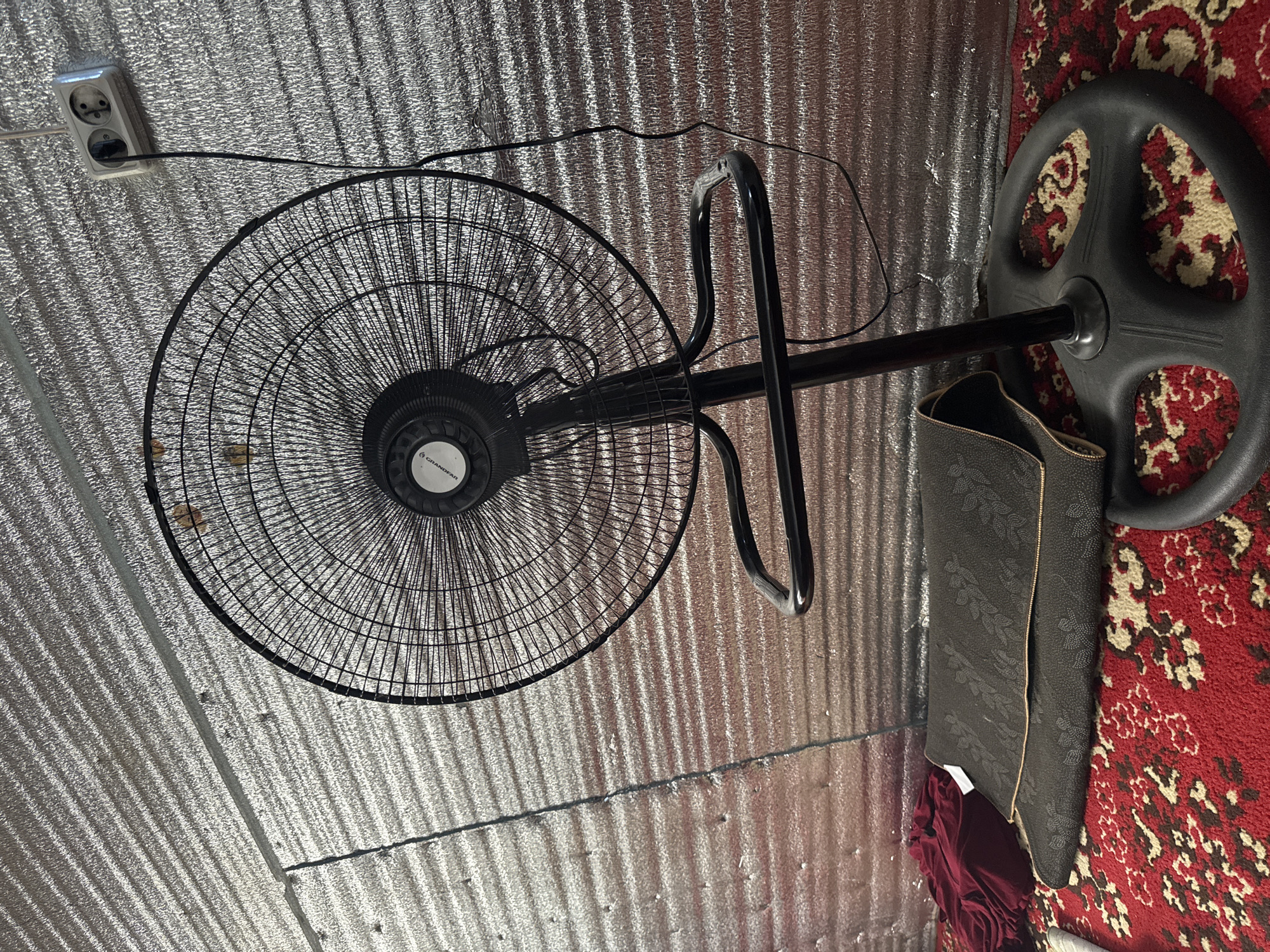 A black oscillating fan stands in a room with a metallic, corrugated wall and a red, patterned carpet. A dark-colored mat is folded in front of the fan. To the left of the mat, a burgundy piece of fabric is visible. A power cord from the fan is plugged into a double electrical outlet mounted on the wall.