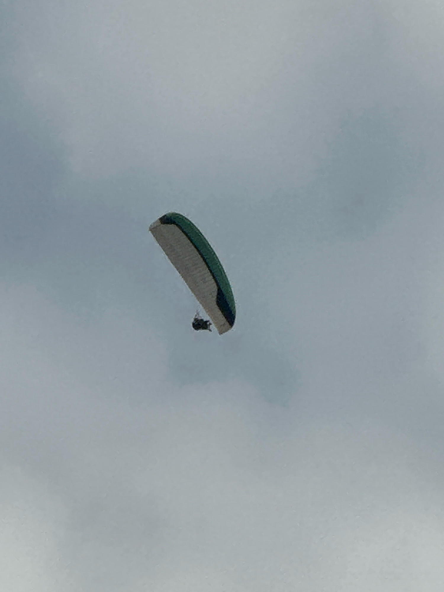 A paraglider is captured in mid-air against a pale, overcast sky. The paraglider, a colorful wing with green and white sections, is angled downwards, with a person suspended beneath it in a dark harness. The scene appears to be an outdoor activity, likely a recreational or sporting event, given the presence of the paraglider. The overall lighting suggests a diffused, possibly cloudy day, with no direct sunlight visible. The image is a medium shot, focusing on the paraglider and its occupant against the vast expanse of the sky. There is no indication of the specific location within Boreti, Montenegro, other than the general context of the sky and paragliding activity.