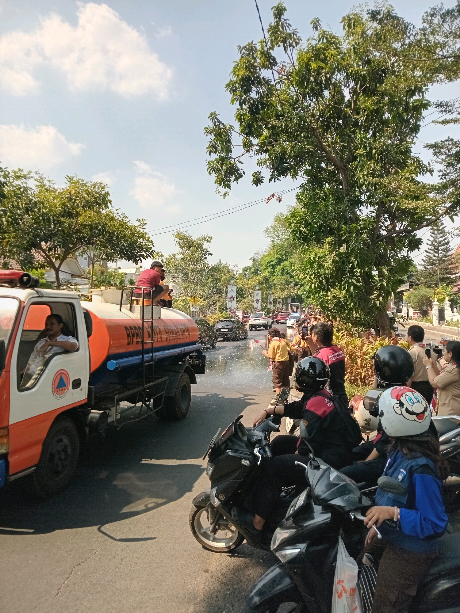 A fire truck is spraying water onto the street in Surakarta, Indonesia. A man is sitting on top of the truck, and another man is driving. Several people are standing on the side of the road, watching the truck. The sky is blue, and there are trees and buildings in the background.