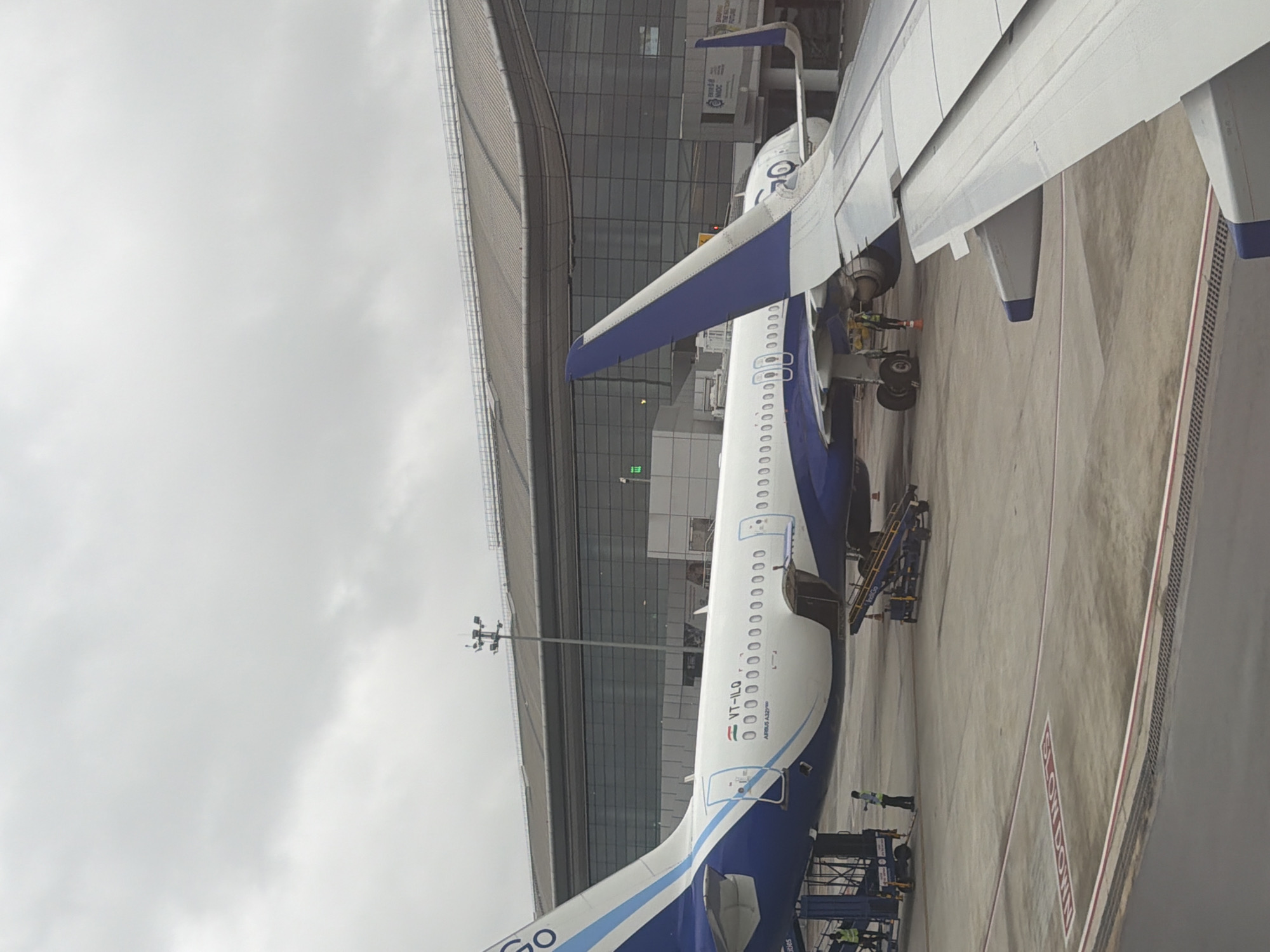The image shows a blue and white Indigo Airlines aircraft parked at an airport, likely Rajiv Gandhi International Airport in Hyderabad, India, based on the presence of Indigo Airlines and the typical architecture of large Indian airports. The aircraft's tail fin, partially obscured by the wing of another aircraft in the foreground, displays the airline's logo. The fuselage is visible, with the registration "VT-ILQ" and "AIRBUS A321neo" clearly marked. Two passenger jet bridges are connected to the aircraft, one on the left and another on the right, indicating boarding or disembarking activities. Ground crew members, one wearing a yellow safety vest, are present near the jet bridges and the aircraft. In the background, a modern airport terminal building with a glass facade is visible. The ground is paved concrete, with a red and white striped line marking a boundary and a "SLOW DOWN" painted message in white. The sky is overcast with gray clouds.