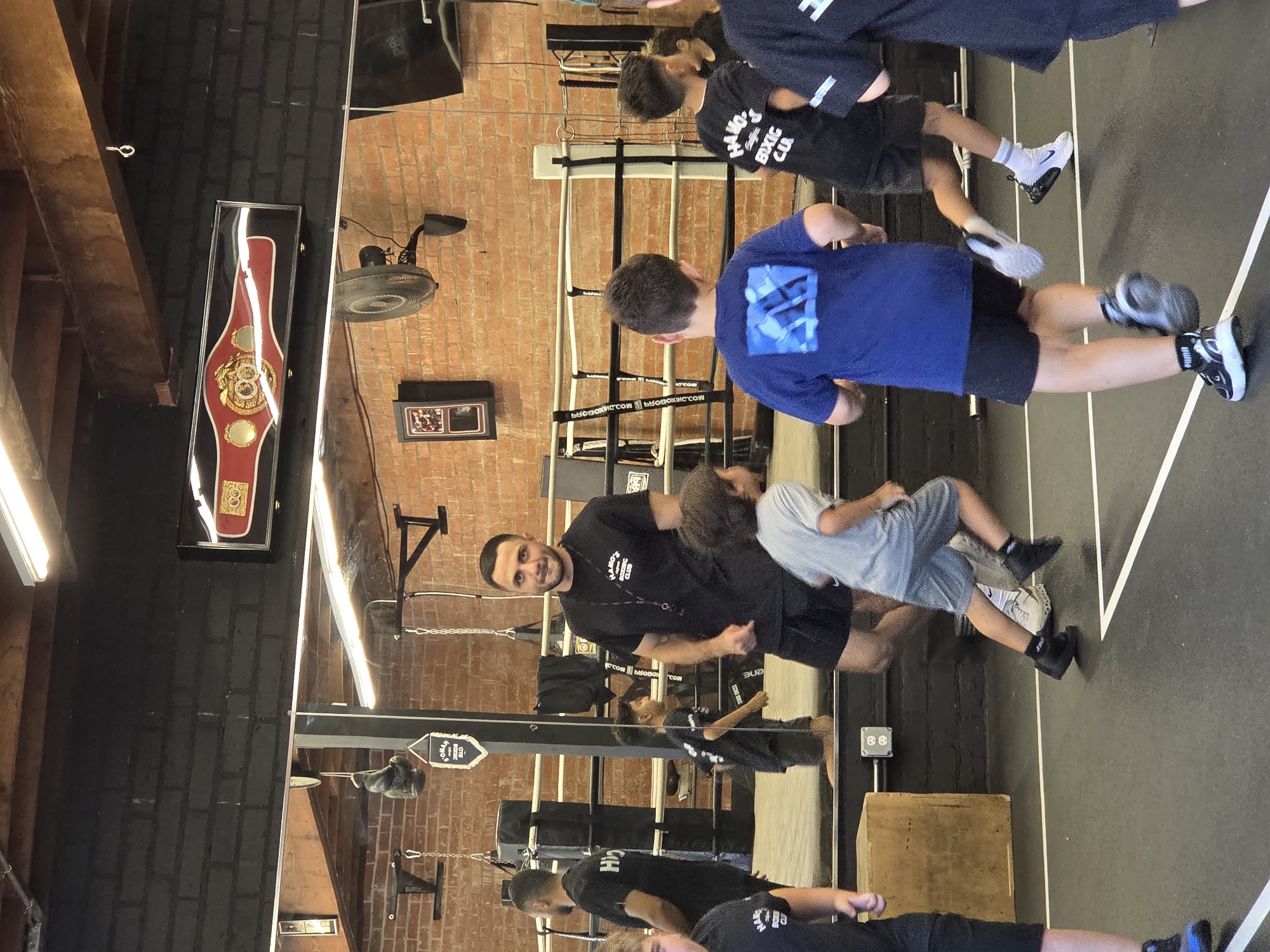 In a boxing gym in Glendale, California, a group of young boys participate in a training session. A male instructor, wearing a black t-shirt and shorts, observes the activity. The gym features a boxing ring, weights, and mirrors. The boys are dressed in athletic wear, and some are running on the mat. A championship belt is displayed on the wall above a mirror, and a fan is mounted on the brick wall. The floor is black with white markings. The overall atmosphere suggests a structured and energetic training environment.