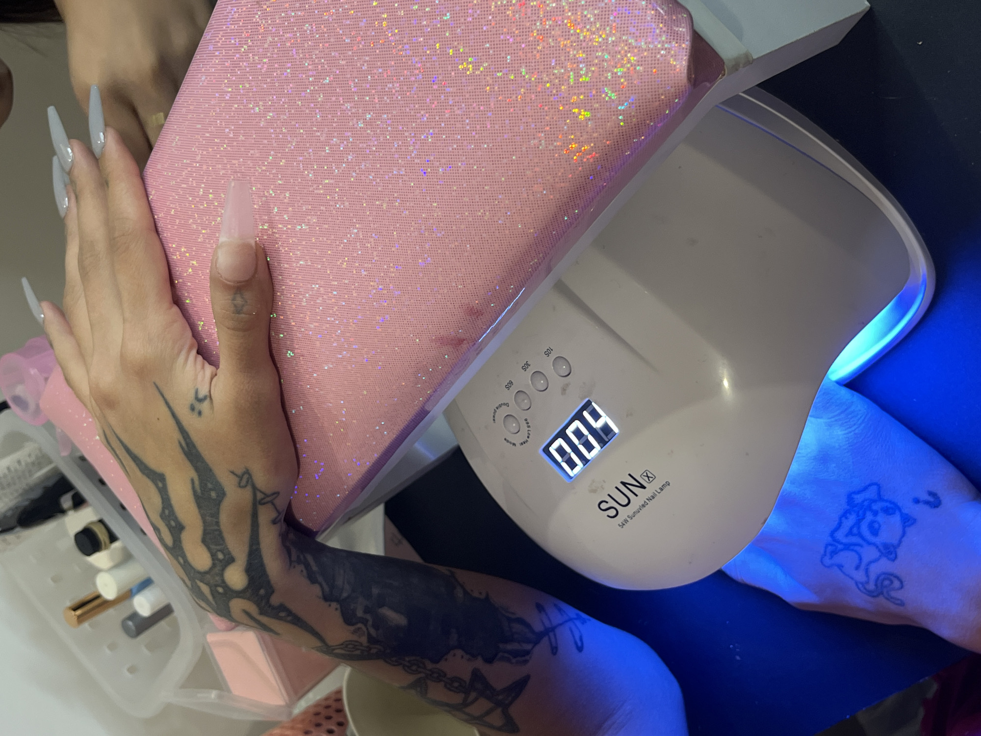 This image, taken in Buenos Aires, Argentina, captures a close-up view of a person engaged in a nail care activity. The primary subjects are the person's hands and forearms, positioned around a white UV nail lamp and a pink, glittery hand rest.
The left hand, adorned with long, pointed, light-colored acrylic nails (except for the thumb which is shorter and squoval), rests on the iridescent pink glitter pillow. The forearm of this hand displays a prominent, dark, flame-like or tribal tattoo extending from the wrist upwards, with a smaller, simpler tattoo visible closer to the knuckles.
The right forearm and hand are placed inside the active UV nail lamp, which emits a bright blue light. A cartoon-style tattoo of a cat with a question mark above its head is visible on the right wrist/forearm. The nail lamp, branded "SUN X" and labeled "54W Sunuvled Nail Lamp," features several buttons for different curing times ("10S", "30S", "60S", "Double power / 99S Low Heat Mode") and a digital display currently showing "004", likely indicating seconds remaining on a timer.
To the left, a clear plastic organizer tray holds various nail care items, including nail polish bottles and tools. A small pink box and a light-colored bowl are also partially visible in the foreground. The background behind the lamp is dark blue/black, contrasting with the lighter elements of the nail station. The scene suggests an indoor setting, likely a home or professional nail salon, where the individual is curing gel or acrylic nails.
