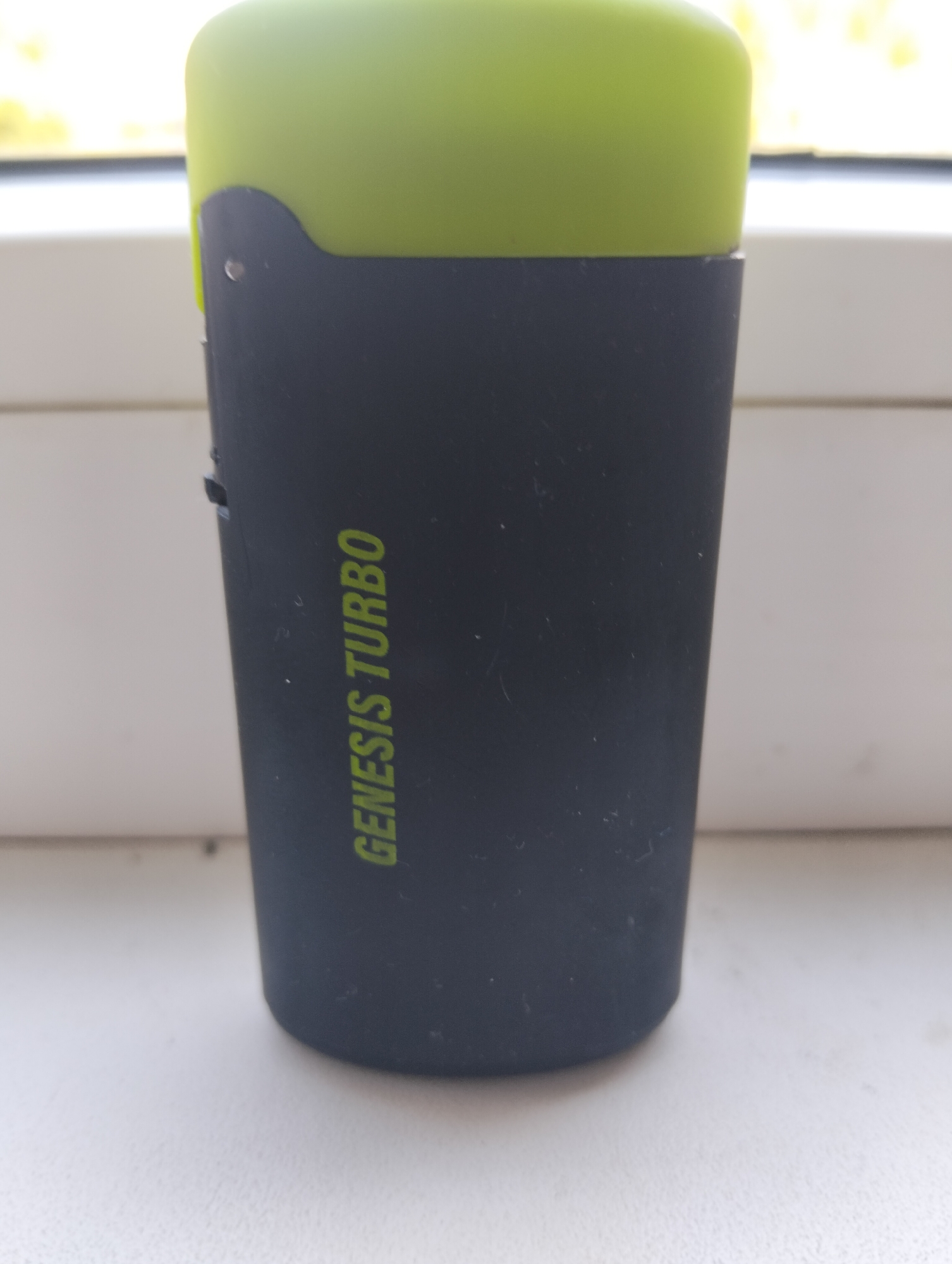 A black cylindrical object with a green top rests on a white surface. The object has the words "GENESIS TURBO" printed vertically in green text on its side. The background is blurred but suggests an indoor setting with a window and natural light. The object appears to be a portable electronic device, possibly a vape pen or a small speaker. The scene is static and shows no people or activity. This image was taken in Cherkessk, Russia.