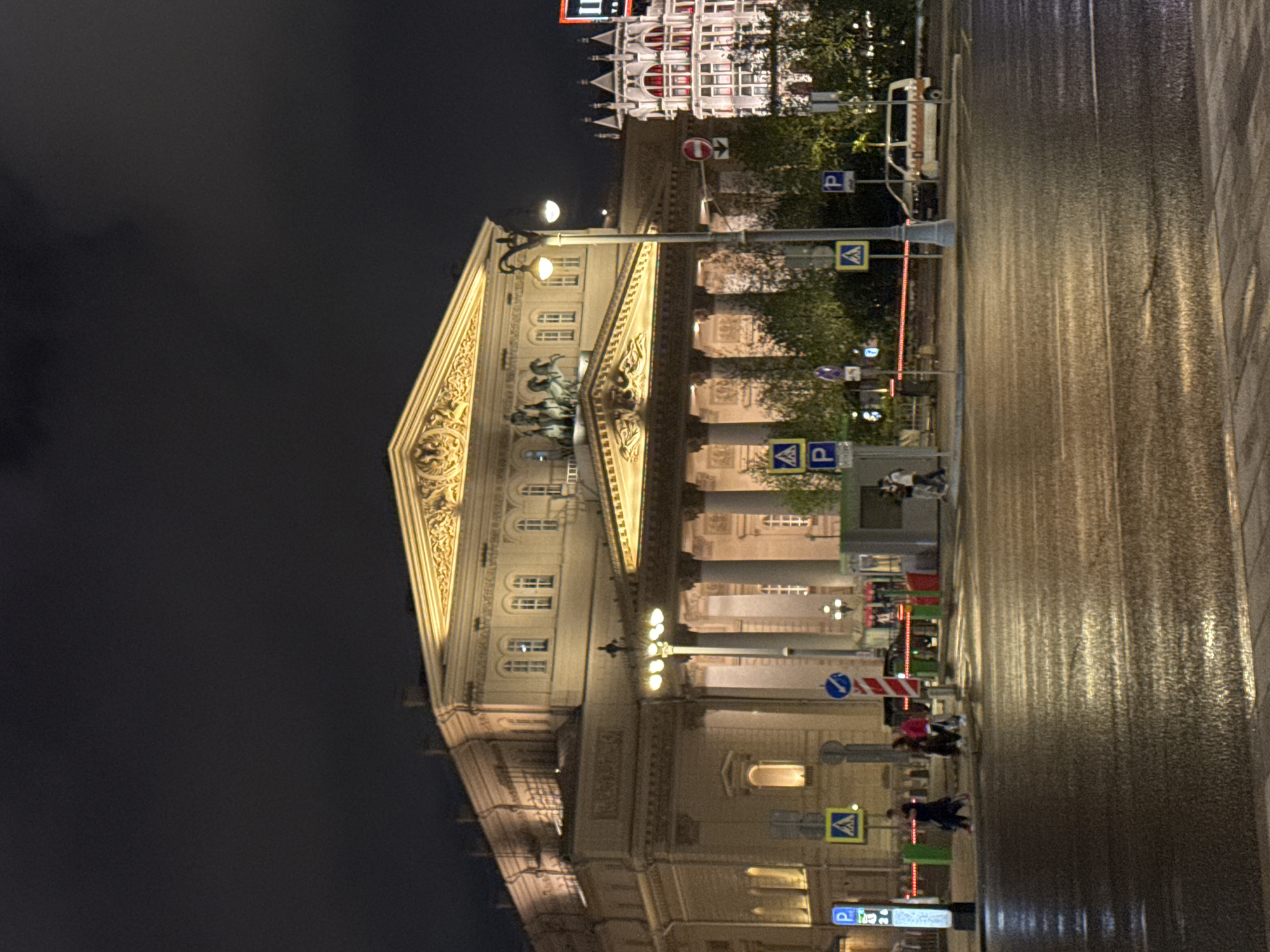 This is a night-time, eye-level, wide-angle photograph of the Bolshoi Theatre in Moscow, Russia. The grand neoclassical building is illuminated, its golden-trimmed facade and colonnade clearly visible against the dark sky. A sculpture of Apollo and his horses adorns the pediment. To the right, a brightly lit, ornate white building with red-trimmed windows stands out. The street in front of the theatre is wet, reflecting the lights, suggesting recent rain. Several traffic signs, including yellow diamond-shaped pedestrian crossing signs and blue parking signs, are present. Pedestrians are walking across the street and on the sidewalk. A white minivan is parked on the street. The overall scene depicts a well-known landmark in Moscow at night.