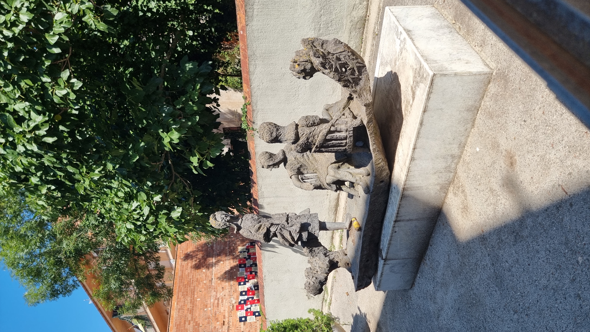 The image depicts an outdoor sculpture located in Olot, Spain. The sculpture is made of stone and features several figures. In the foreground, a long, irregular stone base supports the main sculptural elements. On the left side of the base, a standing figure of a person is depicted, appearing to be dressed in layered clothing. To the right of this figure, two seated figures are positioned on a bench-like structure within the sculpture. One seated figure faces left, while the other faces forward. Both are rendered in a stylized manner. At the far right end of the stone base, another sculpted element resembling an animal or creature is present. The sculpture is situated on a white concrete pedestal. The surrounding environment includes a grey concrete ground, a low, rough-textured concrete wall behind the sculpture, and a brick wall further back adorned with a checkered pattern of red, white, and blue tiles. Lush green trees and foliage are visible above and behind the wall, and a portion of a building with balconies can be seen to the left. The sky is clear and blue. The lighting suggests it is daytime, with shadows cast on the ground.