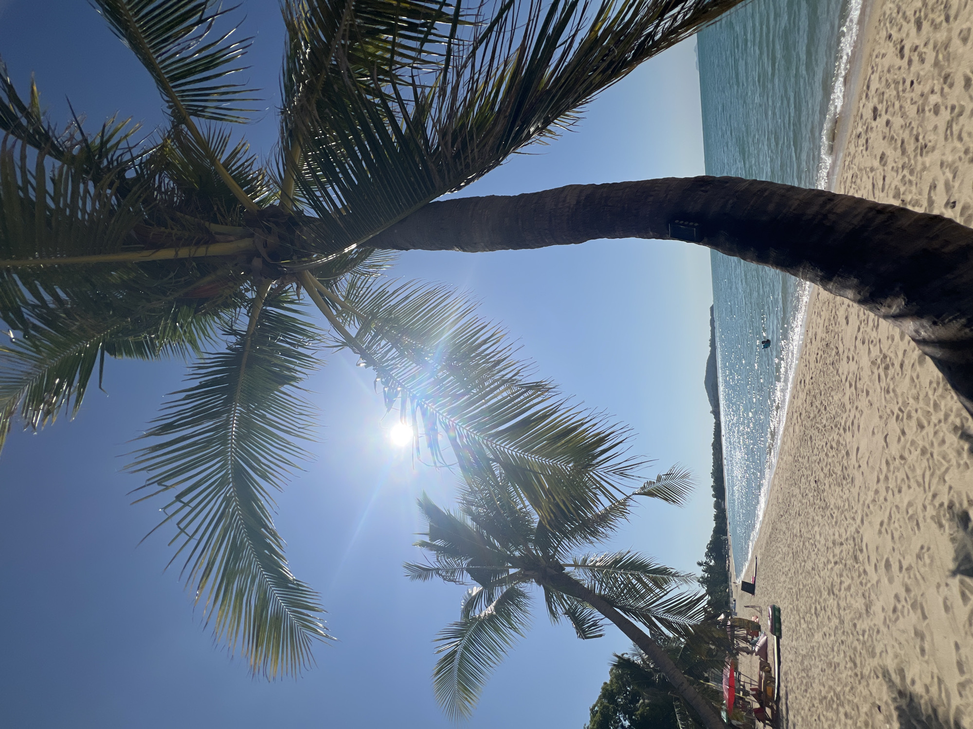 The image depicts a tropical beach scene in the Ko Samui District, Thailand, under a clear blue sky with the sun shining brightly and creating lens flare. Two prominent palm trees, one in the foreground and another to the left, frame the view. The sand on the beach is golden and shows footprints. Along the shoreline, the turquoise ocean water is calm, with small waves gently lapping the shore. The sunlight sparkles on the water's surface. In the distance, across the water, a coastline with greenery and hills can be observed. On the beach, there are a few beach chairs and umbrellas, suggesting a relaxing atmosphere. A lone swimmer is visible in the water. The image captures a serene beach environment characteristic of Ko Samui.