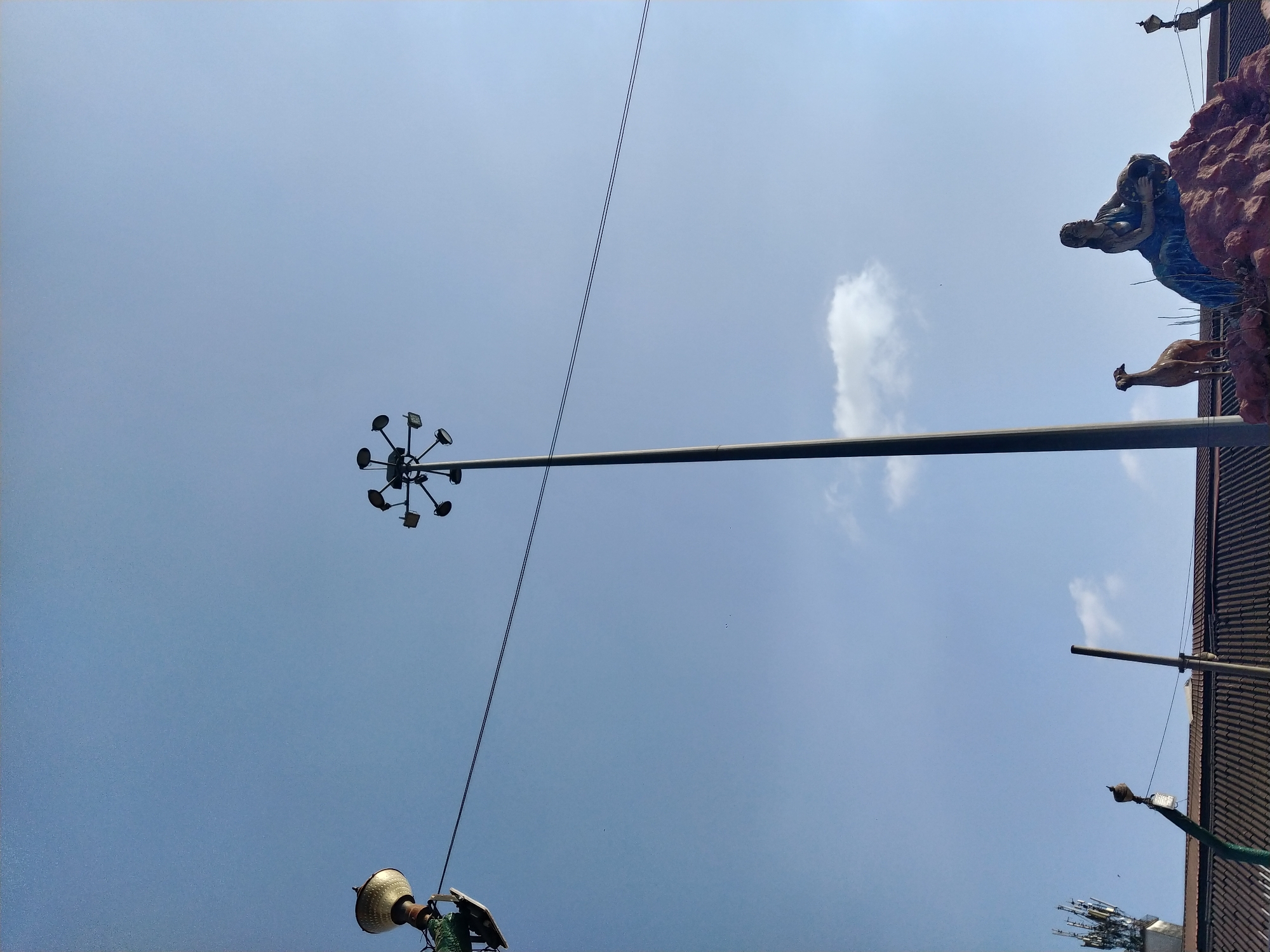 The image, taken from a low angle, shows a view of the sky above Mapusa, India. A tall metal light pole with multiple spotlights extends upwards, intersecting with overhead power lines. To the right of the frame, part of a building is visible, adorned with sculptural elements depicting a woman and a deer. Other smaller light fixtures are attached to the building and a nearby pole. The sky is pale blue with scattered white clouds.