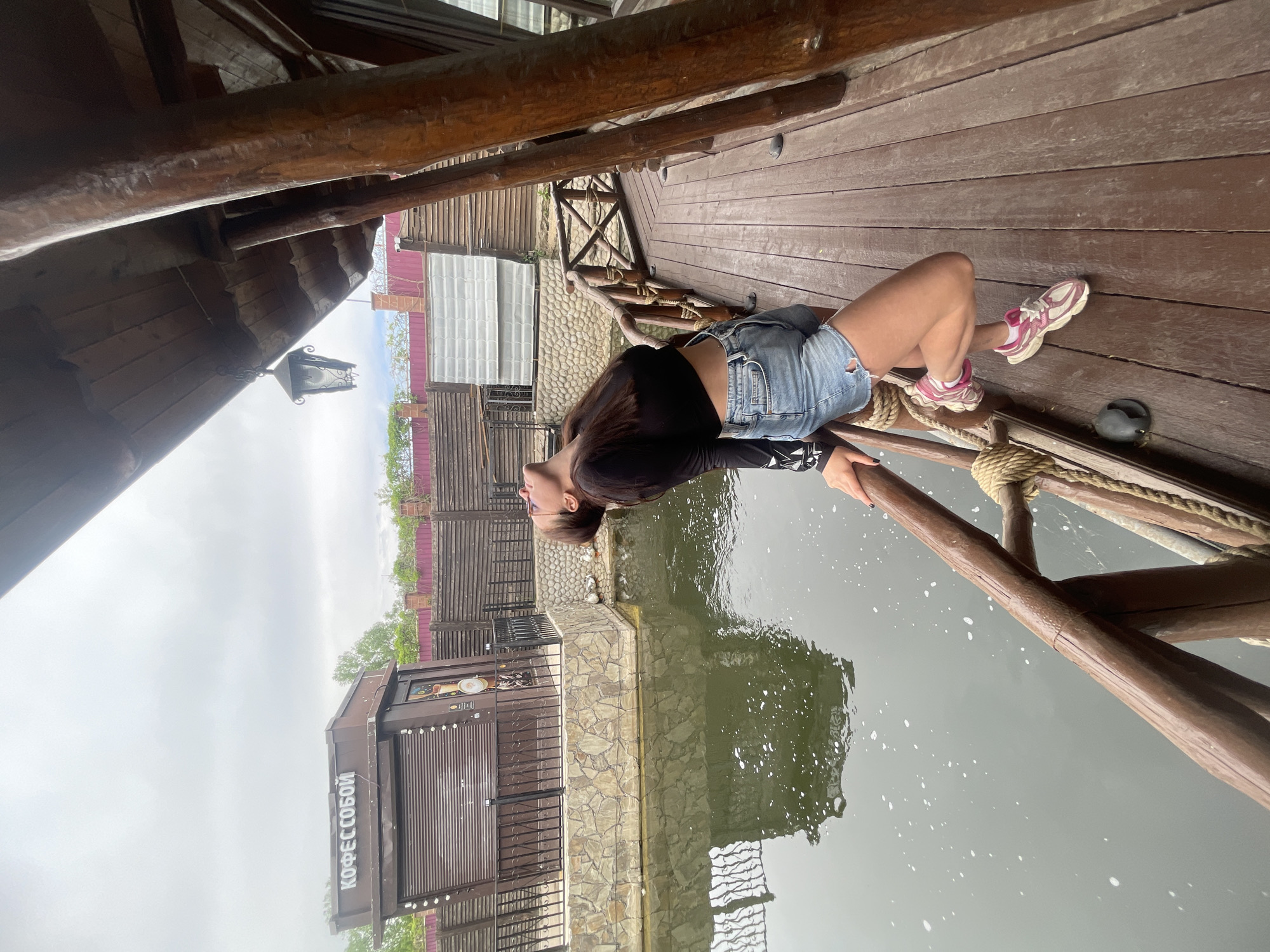 A young woman is lying on a wooden deck by the water's edge. The woman is wearing a black long-sleeved shirt and blue denim shorts. She has on pink and white sneakers. The woman is looking up. There is a wooden structure above the deck. The structure is made of wooden beams. Beyond the deck and water, there is a building with a sign in Russian that says "КОФЕС СОБОЙ" (Coffee to go). The building is brown with a stone base. In the background, there is a fence and some vegetation. The water appears to be a lake or river. The location is Molokovo, Russia.