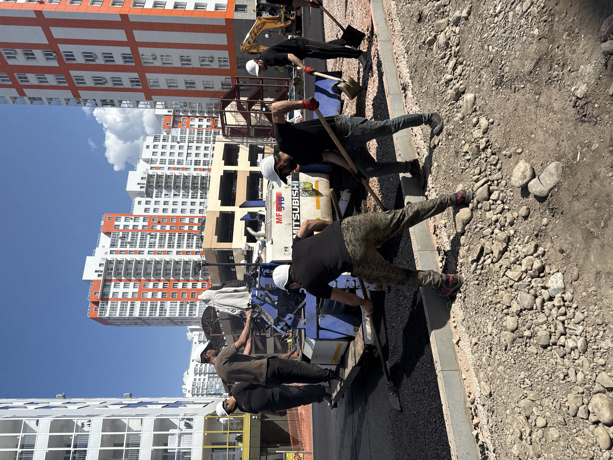 The image shows a construction site in Krasnoyarsk, Russia. Several construction workers are operating an asphalt paving machine on a road. Three workers are near the machine, while others are using shovels to work on the asphalt. They all appear to be wearing hard hats. The paving machine bears the Mitsubishi name. In the background are several tall apartment buildings, one orange and white, and another partially constructed. The sky is clear blue with some clouds. The ground is a mix of paved asphalt and gravel.