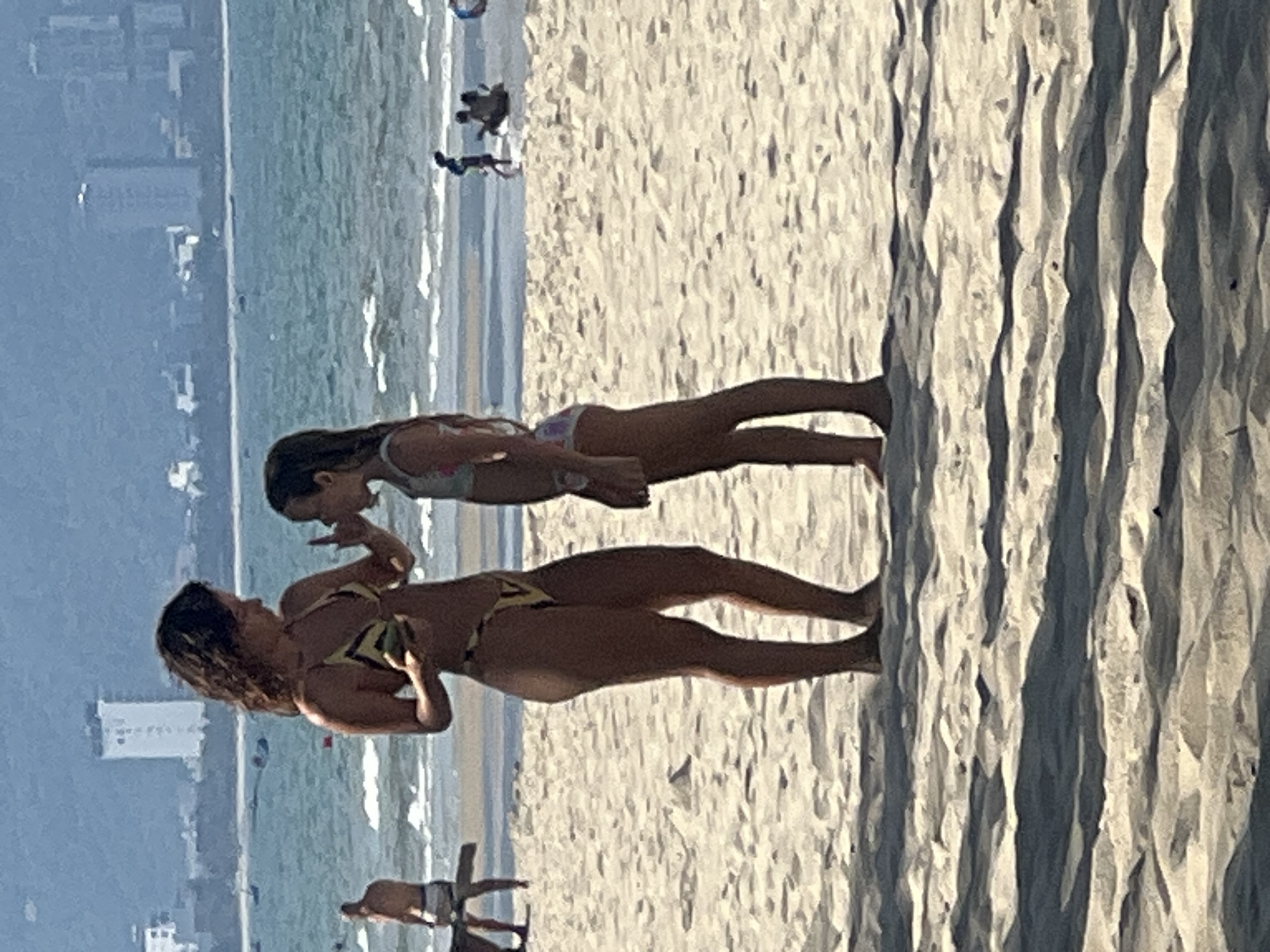 The image captures a sunny day at a beach, likely in Đà Nẵng, Vietnam, with the ocean in the background and the sand in the foreground. Two individuals are the main subjects. The person in the foreground appears to be an adult woman wearing a yellow and black patterned bikini. She is facing left, with her head turned towards the other person. Her arms are raised, and she seems to be in mid-conversation or interaction. Behind her, a younger person, possibly a child, is also visible, wearing a patterned bikini. This younger person is facing the adult and is looking down. The background shows a hazy cityscape with buildings, indicative of a coastal urban area. The ocean is calm with gentle waves. The sand in the foreground is textured, with visible ripples and shadows suggesting bright sunlight, possibly late morning or early afternoon. There are other faint figures in the distance on the beach, suggesting other beachgoers. The overall atmosphere appears relaxed and leisurely, typical of a beach day. No text is visible in the image.