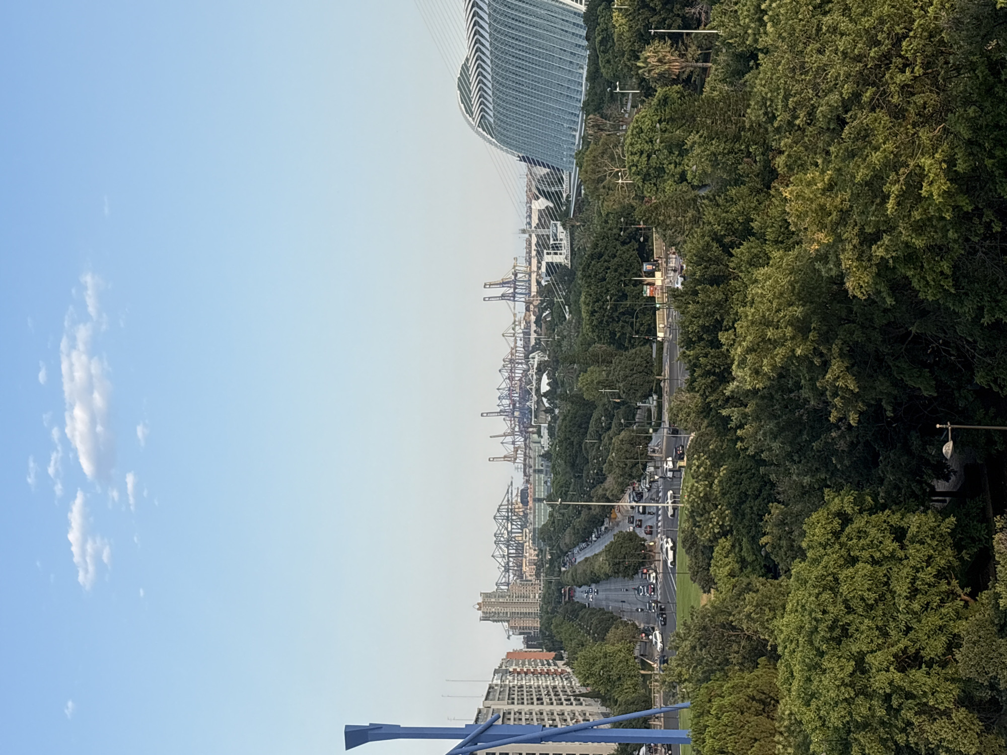 The image captures an expansive, wide-angle view of a bustling port city, likely València, Spain, oriented vertically. The scene is divided into three distinct horizontal bands: a clear, pale blue sky with scattered white clouds occupies the top third on the left, gradually transitioning to a lighter hue on the right.
The middle band showcases a dynamic urban landscape. Dominating the left side of this band is a strikingly modern, curvilinear building with a glass facade and white cable supports, suggesting a significant architectural landmark. To its right, a dense array of industrial port cranes, rendered in shades of gray and red, are visible in the distance, indicating a large, active shipping and logistics hub. A busy road, lined with trees and streetlights, runs through the center of the image, filled with numerous cars, suggesting it's a period of moderate to heavy traffic. Further down this road, several multi-story apartment buildings are nestled among lush green trees, adding a residential element to the urban sprawl.
The bottom band is characterized by the dense foliage of trees, with their leaves casting shadows and creating a textured canopy. Some of the buildings are more visible here, with their architectural details partially obscured by the greenery. A distinctive abstract blue sculpture is partially visible in the lower left corner.
The lighting suggests it is late afternoon or early evening, with the sun casting a soft glow across the scene. The weather appears clear and pleasant. The overall impression is one of a vibrant, modern city with a strong industrial presence alongside residential areas and green spaces. No visible text is present in the image.