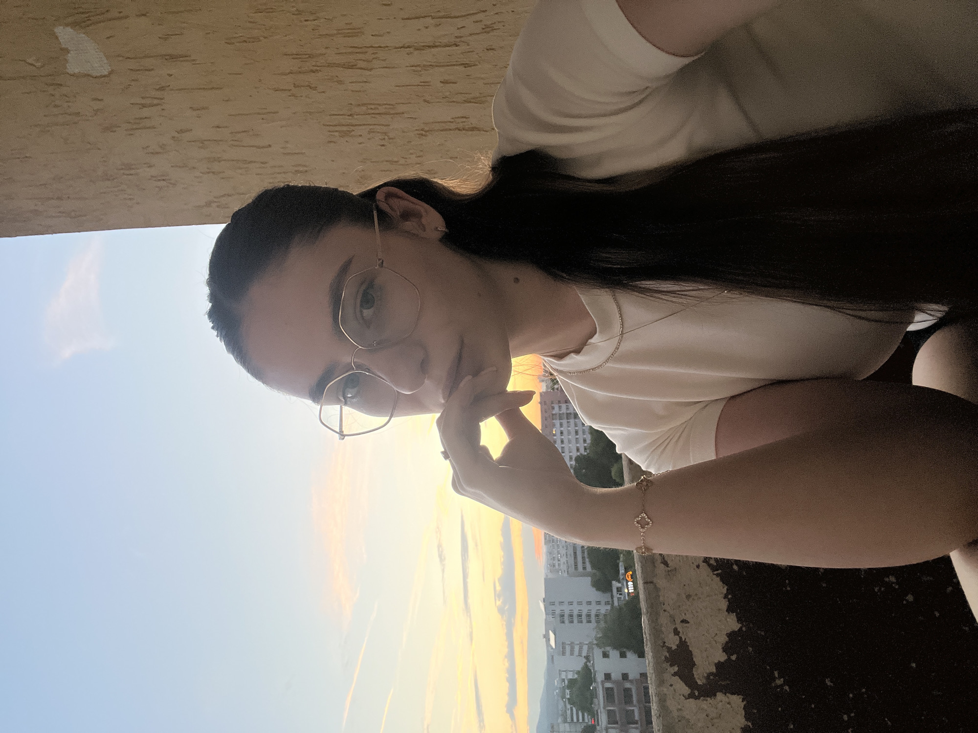 A young woman with dark hair pulled back in a ponytail, wearing a white t-shirt and gold-rimmed glasses, poses for a selfie on a balcony with a city skyline in the background at sunset. She rests her chin on her hand, wearing a bracelet with a clover-shaped charm and a thin necklace. The sky displays a gradient of orange, yellow, and blue hues, with scattered clouds. In the distance, apartment buildings and a faint mountain range are visible. A textured, beige wall is to the right of the woman.