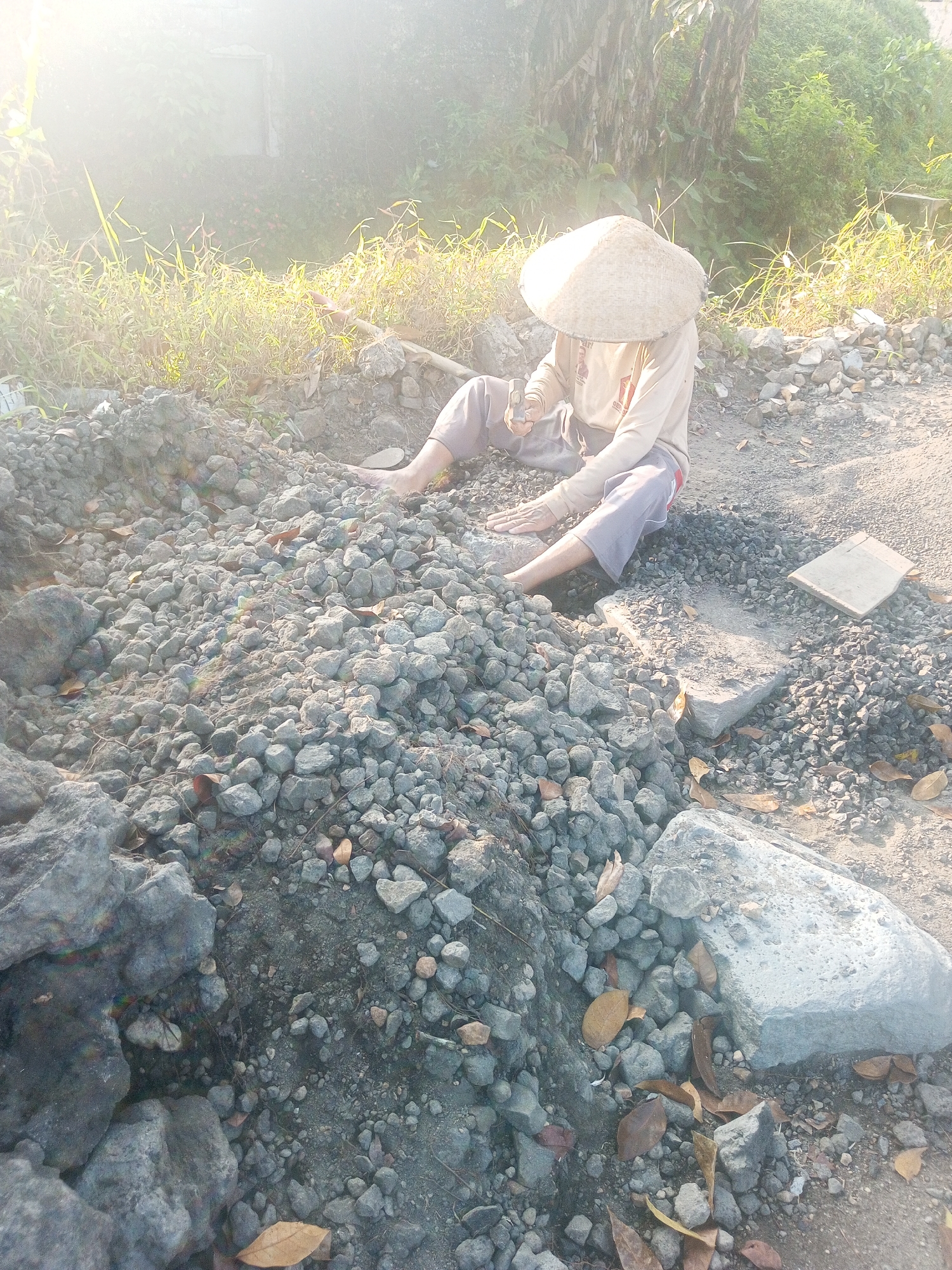In Garung, Indonesia, a person wearing a conical hat and a long-sleeved shirt sits amongst a pile of rocks and gravel. The person's legs are visible, and they appear to be holding a small object in their hands. The ground is covered with various sizes of stones, dirt, and scattered dry leaves. In the background, vegetation and trees can be seen, bathed in bright sunlight that creates a lens flare effect. A white wall is partially visible in the upper left of the image. The overall scene suggests an outdoor work environment, likely a construction or landscaping site, under a clear, sunny sky.