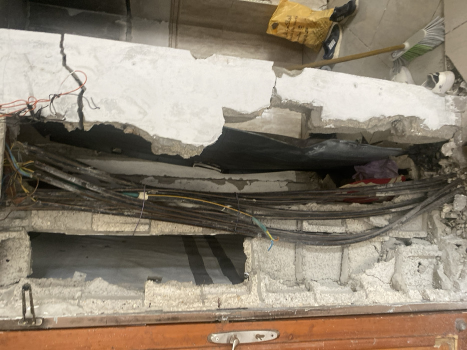 The image displays a severely damaged interior wall section within a building in Khan Yunis, Palestinian Territory. On the left, a partially visible brown wooden door with a silver handle is set within a concrete block wall, which exhibits crumbling plaster and exposed blocks. Adjacent to the door, a vertical cavity reveals a dense bundle of large-gauge electrical cables running upwards, intertwined with thinner, multicolored wires, some with exposed conductors. The wall structure surrounding these cables consists of broken concrete blocks and dislodged plaster. To the right, a vertical section of a white-plastered wall shows extensive damage, with large fragments of plaster missing and a prominent horizontal crack. The floor in the lower right foreground is tiled, and several items are present: a yellow plastic shopping bag with Arabic text, multiple pairs of shoes (including dark and white sneakers), and a green-and-white broom leaning against the damaged wall. No individuals are depicted, and no specific actions or interactions are occurring. The environment indicates significant structural damage or disrepair.