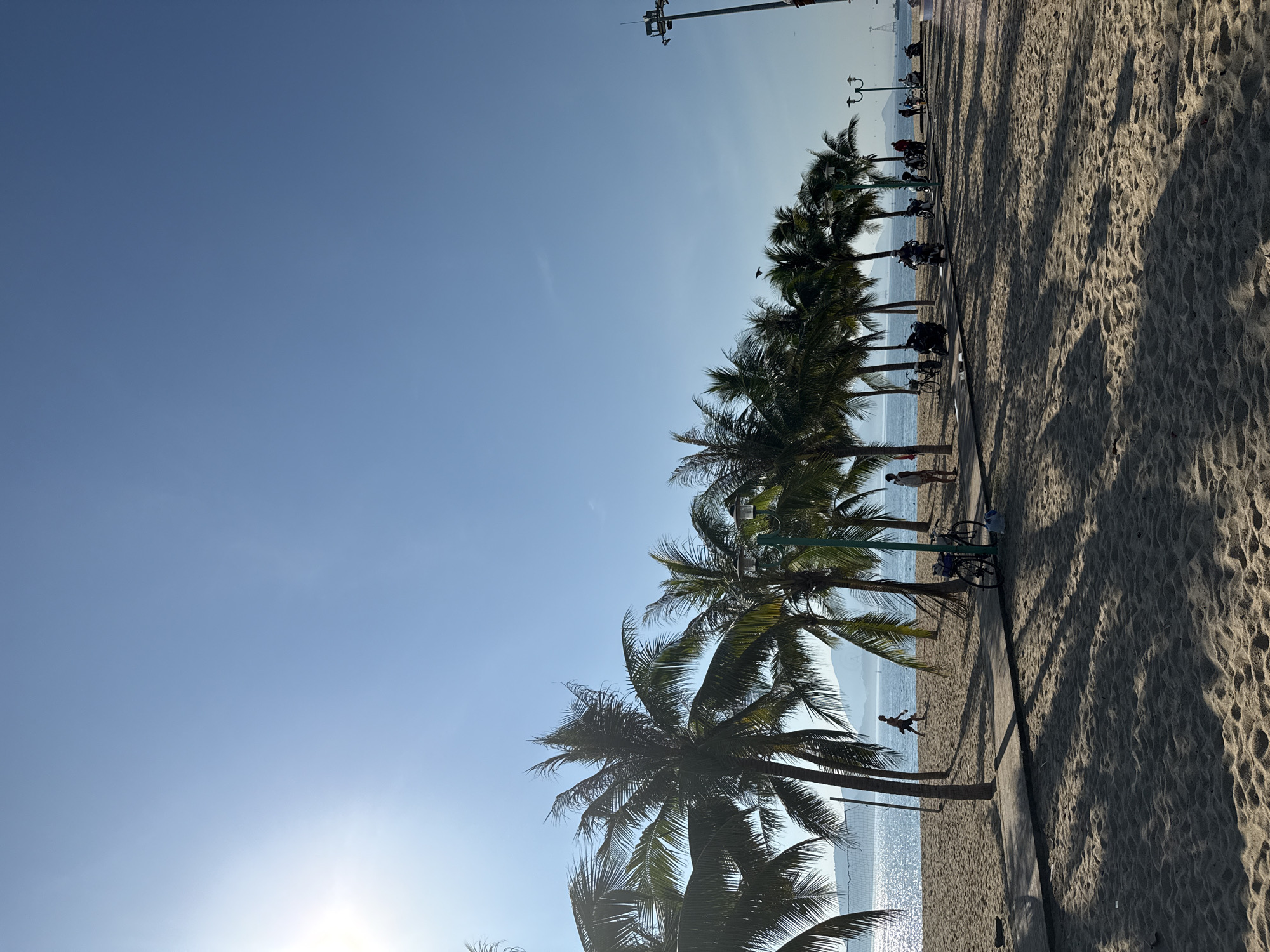 The image depicts a sunny day at a beach in Nha Trang, Vietnam, likely along the Trần Phú Boulevard due to the presence of palm trees, a paved walkway, and the ocean. In the foreground, a sandy beach is visible with sand ripples. A paved pathway runs parallel to the beach, lined with palm trees casting shadows. Several people are present: one person is walking on the sand near a volleyball net, another is standing on the walkway, and a group of people are near parked motorcycles. A bicycle is also parked on the walkway. In the background, the ocean shimmers under the bright sun, with distant islands or mountains visible. The sky is clear and blue, with the sun positioned in the upper left quadrant of the image.
