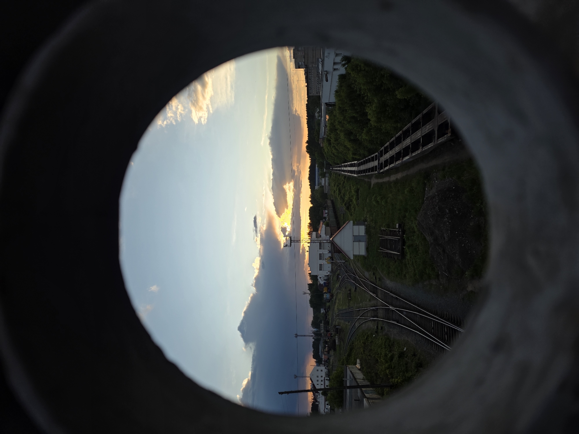 The image shows a sunset scene viewed through a concrete pipe. The sky has a mix of blue and dark clouds, with the sun setting on the horizon, creating a warm, golden glow. Below the horizon, the view encompasses a railway yard with several tracks converging and diverging. There are a few buildings visible among the tracks, and along the right side of the image is a conveyor structure running along the edge of a grassy area with trees. The photograph has been rotated clockwise. According to geolocation data, this photo was taken in the city of Chaikovsky, Perm Krai, Russia.