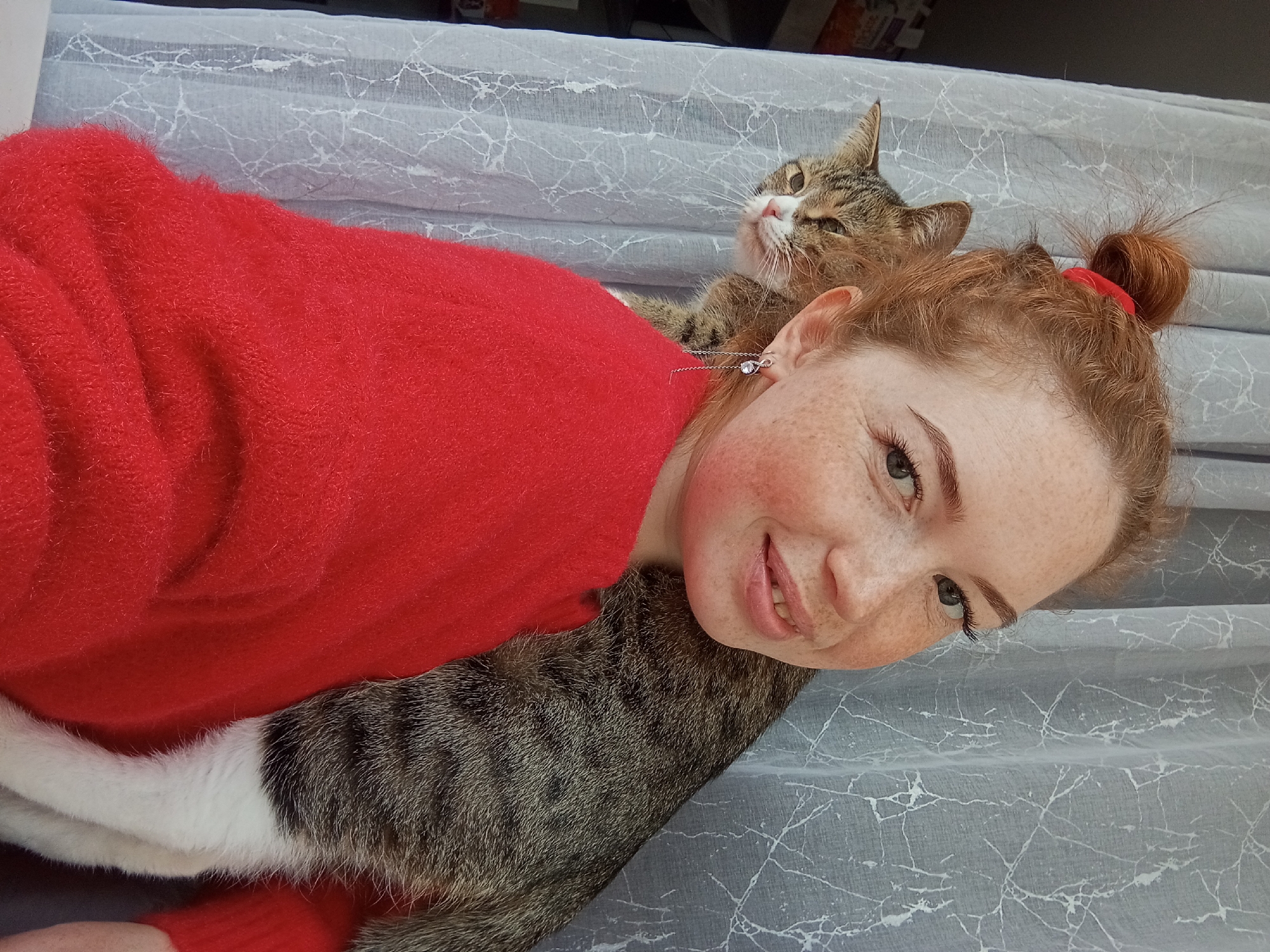 A person with red hair, styled in a bun and secured with a red band, is lying on a light grey surface with a marble-like pattern. The person is wearing a bright red sweater. A tabby cat rests partially on the person's back. The cat's tail and part of its body are visible. The person is looking directly at the camera. The location is unknown, with no visible indication that the scene is in the city of Yurgamysh, Russia.