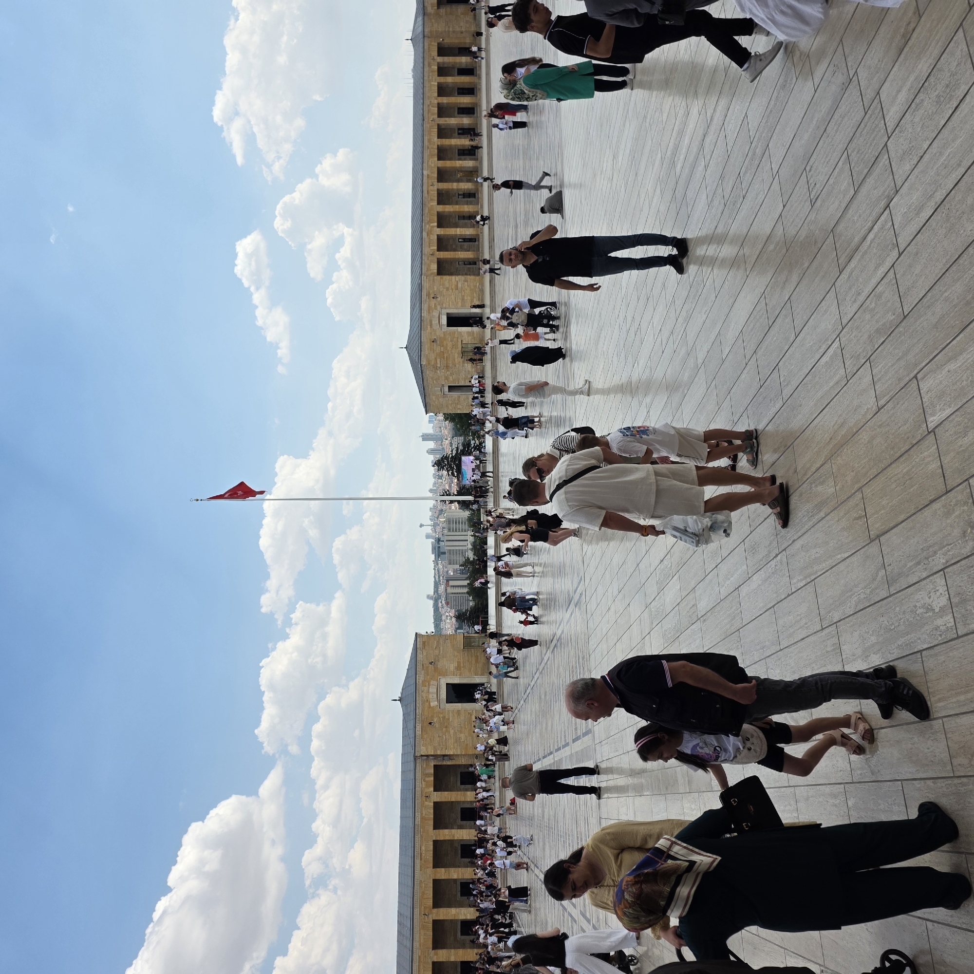 The image depicts a sunny day with scattered clouds at Anıtkabir, the mausoleum of Mustafa Kemal Atatürk, located in Ankara, Turkey. A large, open plaza paved with light-colored tiles dominates the foreground. In the background, two long, symmetrical stone buildings with arched entrances flank the plaza. A tall flagpole stands centrally in the distance, flying the Turkish flag. Numerous people are spread across the plaza, walking, standing, and interacting. Some individuals are dressed in casual summer clothing, while others wear headscarves or traditional attire. The scene suggests a public space with a mix of tourists and locals visiting the historical site.