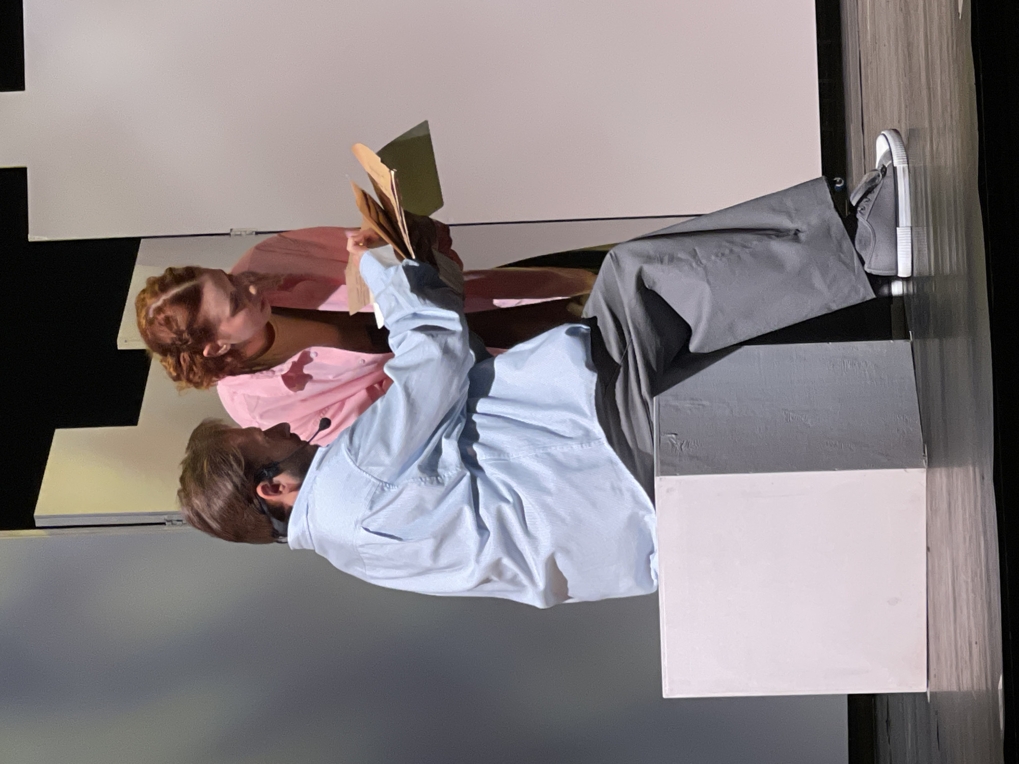 Two people are on a stage. One person, wearing grey pants, grey shoes, and a light blue shirt, is sitting on a grey and white structure. The other person, wearing a pink shirt, is next to the first person. Both are looking at papers being held by the first person. The background consists of white and black planes. The location is not indicated.