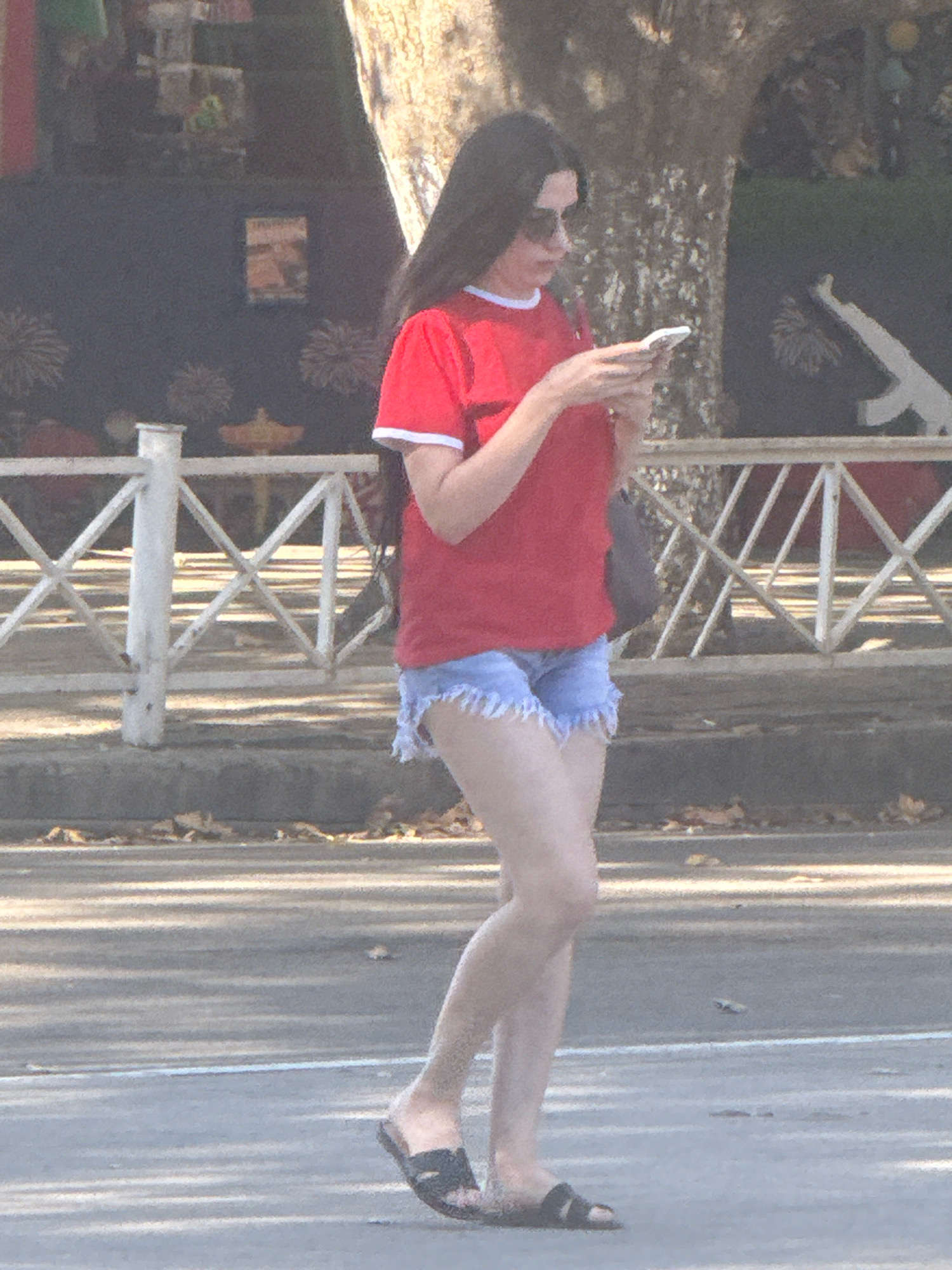 A full-body shot, taken from the side, shows a young woman walking and looking at her phone. She has long dark hair and is wearing sunglasses, a red t-shirt with white trim, and denim shorts. She is also wearing black sandals. The woman is walking on a paved surface, likely a sidewalk or promenade, with a tree-lined area behind her and a white metal fence. The background appears to be a public space with some foliage. There are some indistinct objects in the background, possibly kiosks or stalls. The lighting suggests it is daytime with some dappled sunlight. The image quality is somewhat grainy and the colors are muted. The scene is likely outdoors in a city environment.