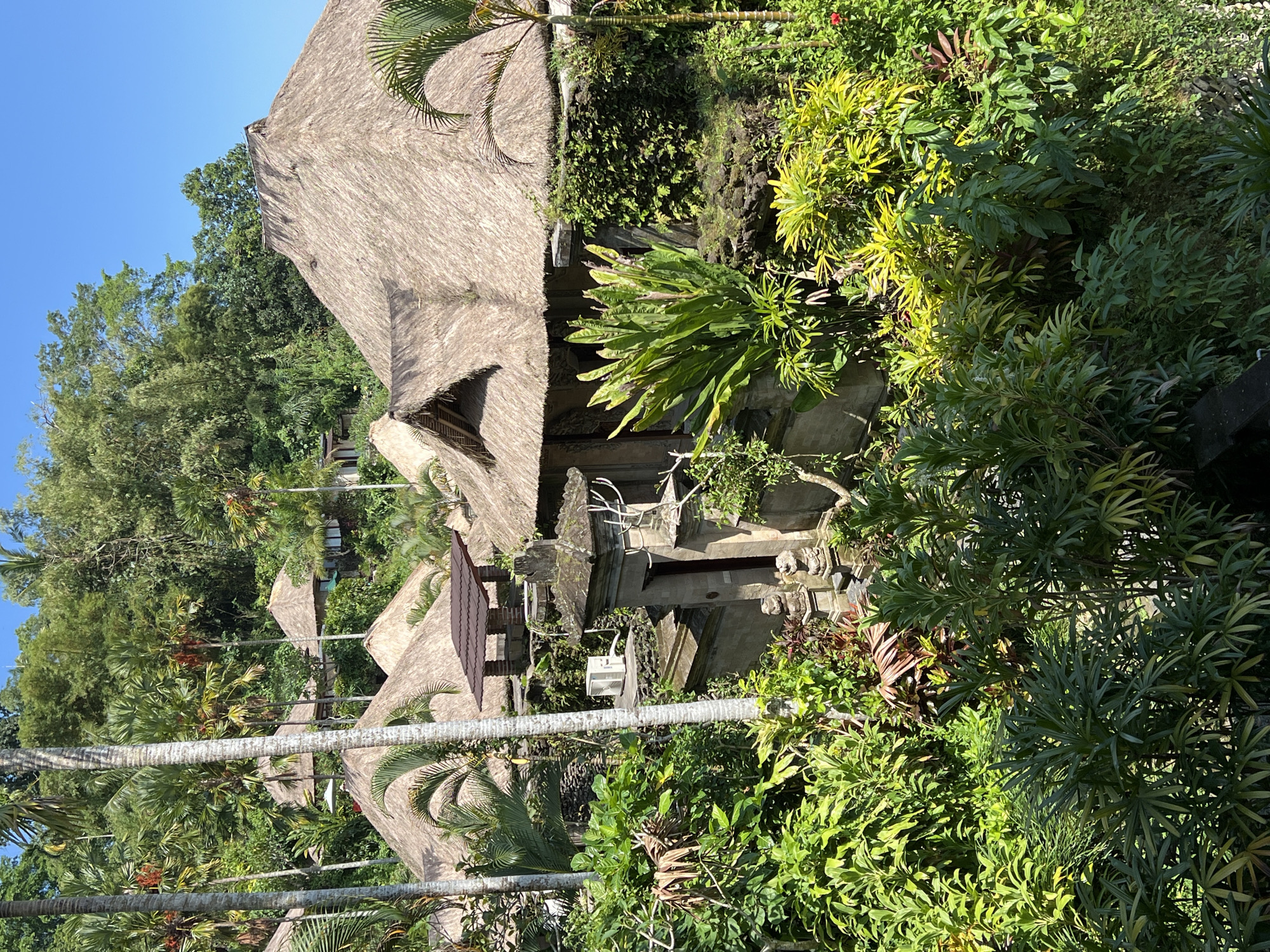 The image captures a scene in Kedewatan, Indonesia, featuring traditional thatched-roof buildings amidst lush tropical vegetation. The structures appear to be part of a resort or residential complex, with multiple buildings visible at varying distances. The foreground is dominated by dense greenery, including various shrubs, trees, and palm fronds. A prominent stone structure or gate, possibly part of a temple or entrance, is nestled among the vegetation. The sky is clear and blue, indicating a sunny day. The overall impression is one of tranquility and integration of traditional architecture with the natural environment.