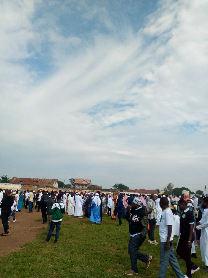 A large gathering of people are assembled in a field in Beni, Democratic Republic of Congo, located at coordinates 0.489 latitude and 29.463 longitude. The field is covered in short green grass. The sky is mostly cloudy with some areas of blue sky visible. The people are dressed in a variety of clothing, including traditional African garb and western attire. There are many people wearing white robes and headscarves. Many people are looking around and interacting with each other. There are some buildings visible in the background, including a one-story building with a yellow sign. Some people are walking away from the camera, and some are walking towards the camera. There is a man wearing a green jacket and black pants walking to the left of the image, and a man wearing a black jacket and black pants walking to the right of the image. There are many trees in the background. There is a small group of people gathered in the lower right corner of the image. The atmosphere appears peaceful and celebratory.