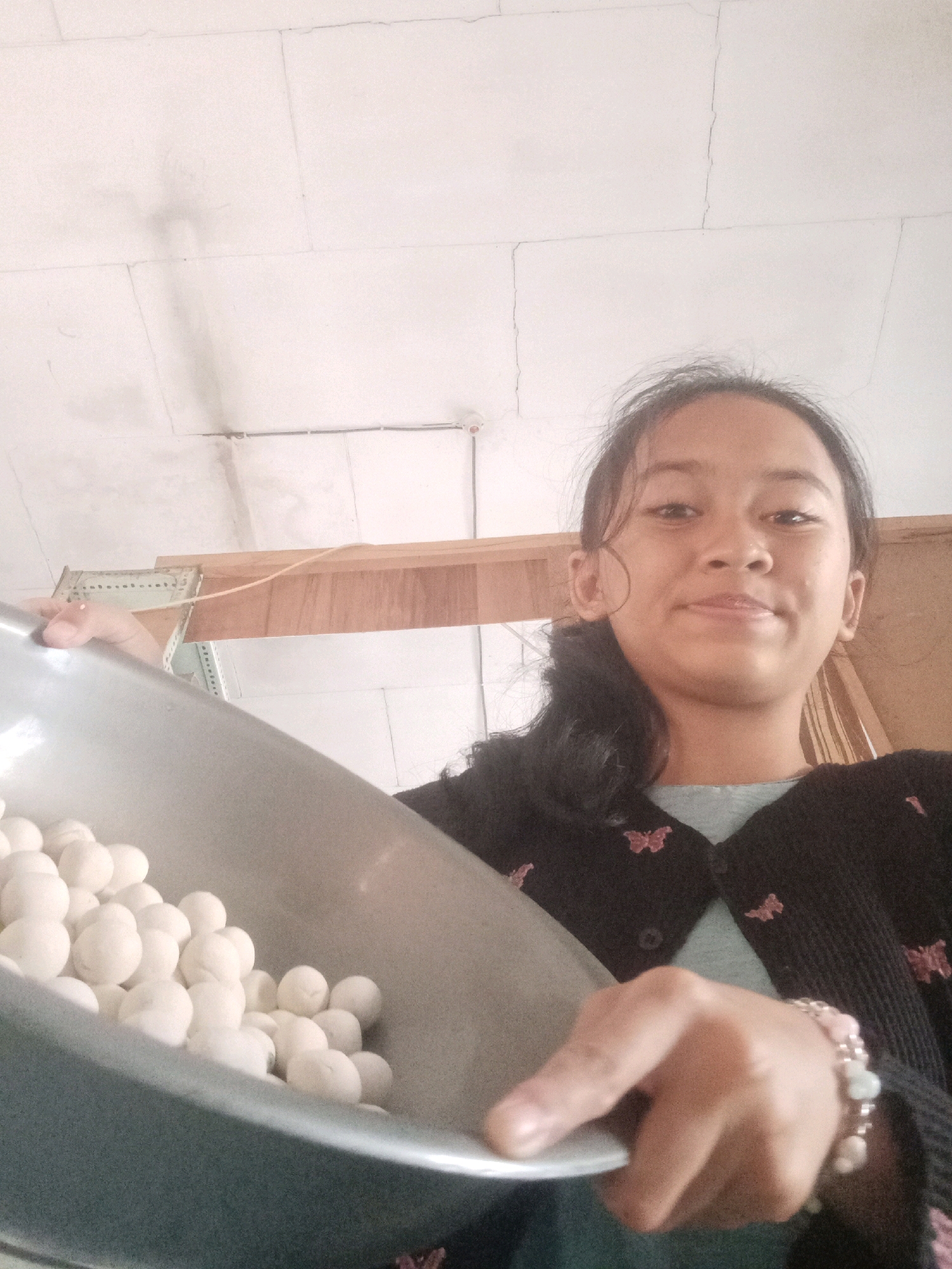 A young Indonesian girl with dark hair pulled back into a ponytail is in the foreground, holding a large, gray metal bowl filled with numerous small, white, round objects, likely food items. She is wearing a dark cardigan with pink butterfly embroidery over a light blue shirt, and a bracelet with pink and white beads on her right wrist. The background appears to be an indoor domestic setting in Badas, Indonesia, with a white ceiling showing cracks and water stains, and wooden shelving with some electrical wiring visible. The lighting suggests an indoor environment, possibly during the daytime.