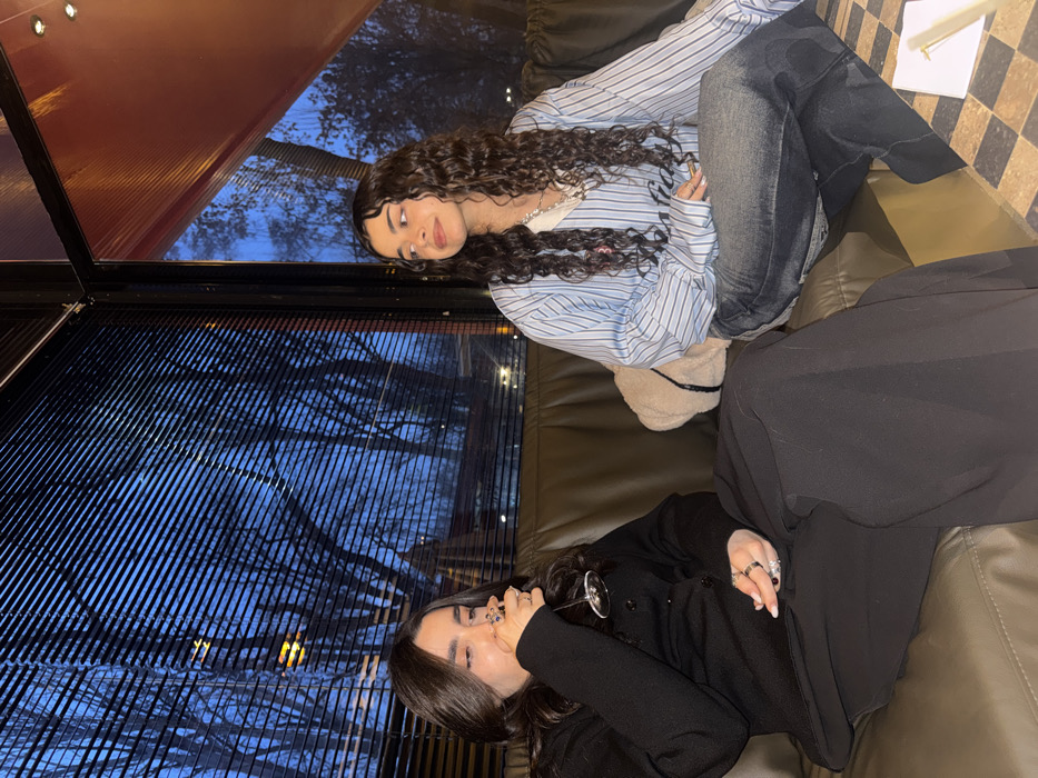 Two young adult females are seated inside a modern cafe or restaurant in Yerevan, Armenia, during twilight hours. The woman on the left, with long dark hair, is dressed in a black cardigan and black wide-leg pants. She holds a stemmed glass containing a dark liquid to her mouth, appearing to drink, and wears multiple rings on her left hand. To her right, the second woman, with long dark curly hair, wears a light blue and white striped long-sleeved shirt and faded dark jeans. She is looking to her right with a slight smile. A light-colored, possibly sherpa, small bag or cushion rests behind her on the dark brown booth seating.
The interior features continuous dark brown (likely leather or faux leather) booth seating. Large, dark-framed corner windows dominate the background, with horizontal blinds covering the upper left pane. Through the windows, bare deciduous trees are visible against a deep blue, twilight sky, indicating a view of a park or a tree-lined area. Faint outdoor lights are discernable in the distance. An internal architectural element in the upper right consists of a reddish, possibly translucent and corrugated, ceiling panel. A checkered table surface (black and brown squares) with a small white object is visible in the bottom right foreground. The specific architectural style, including the windows with blinds and the reddish ceiling element, aligns with establishments such as **The Green Bean** cafe, particularly locations offering a park view, like those near **Tumanyan Park** or along **Isahakyan Street** in the vicinity of the **Cascade Complex** in Yerevan.