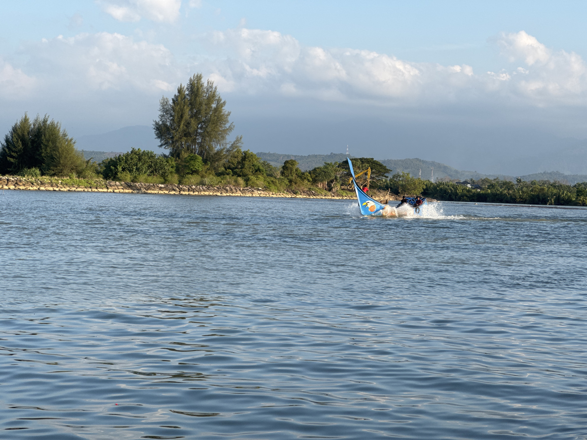 In the city of Meunasah Jurong, Indonesia, a traditional Indonesian fishing boat, adorned with a blue and yellow sail featuring an eagle motif, cuts through the tranquil blue water. The boat, propelled by an outboard motor, creates a white spray as it moves. Two individuals are visible on the boat, one steering and the other seated. The boat is positioned in the foreground, leaving a wake behind it. In the mid-ground, a rocky embankment lines the shore, topped with lush green vegetation and tall trees, including pine trees. Beyond the shoreline, a range of hills, partially obscured by mist and clouds, forms the backdrop. The sky above is a pale blue, dotted with fluffy white clouds. The overall scene captures a moment of activity on the water with a natural landscape.